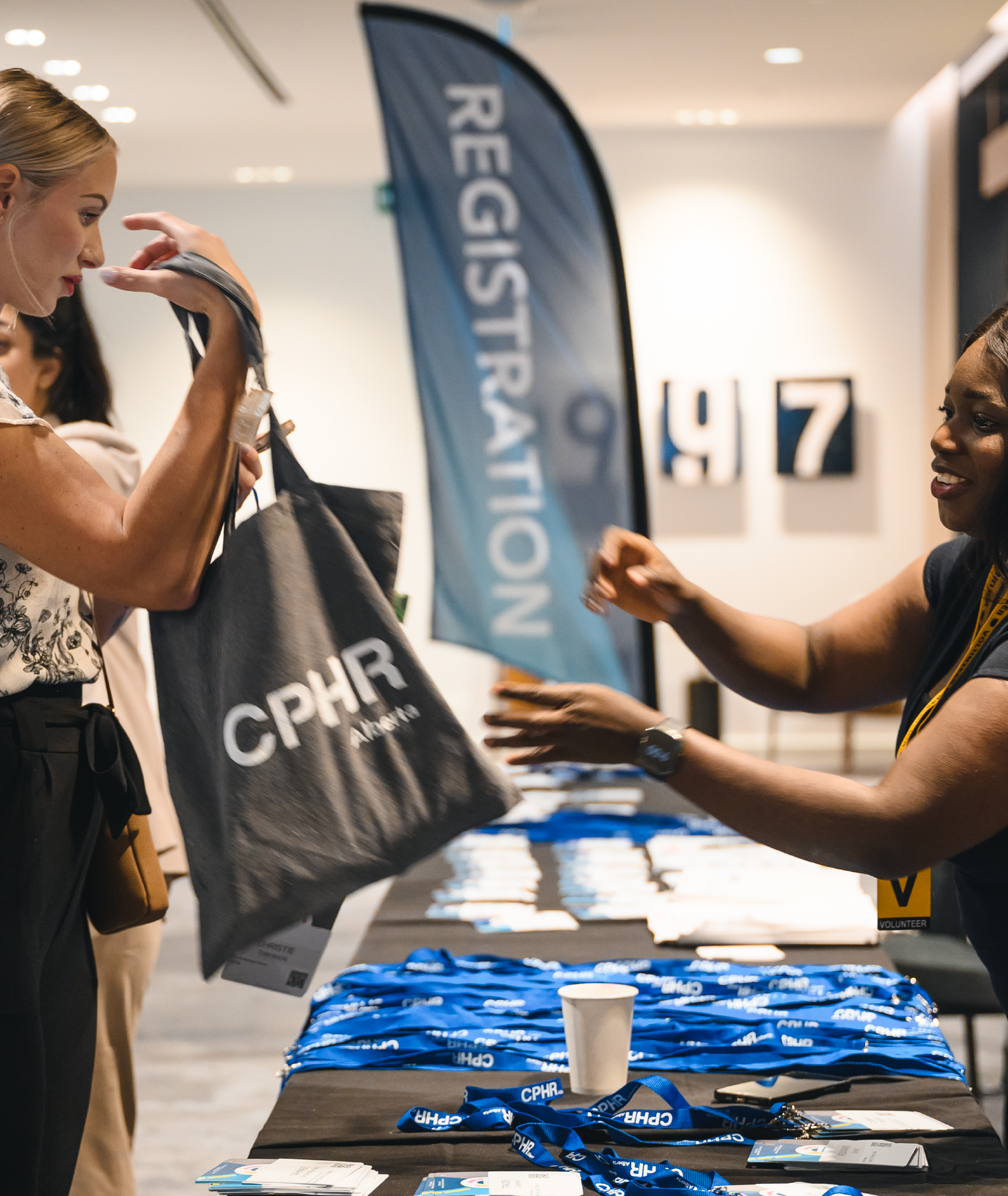 Two men in conversation. One with a beard, glasses, and a blue sweater. The other wearing glasses, holding papers, and a patterned blazer.