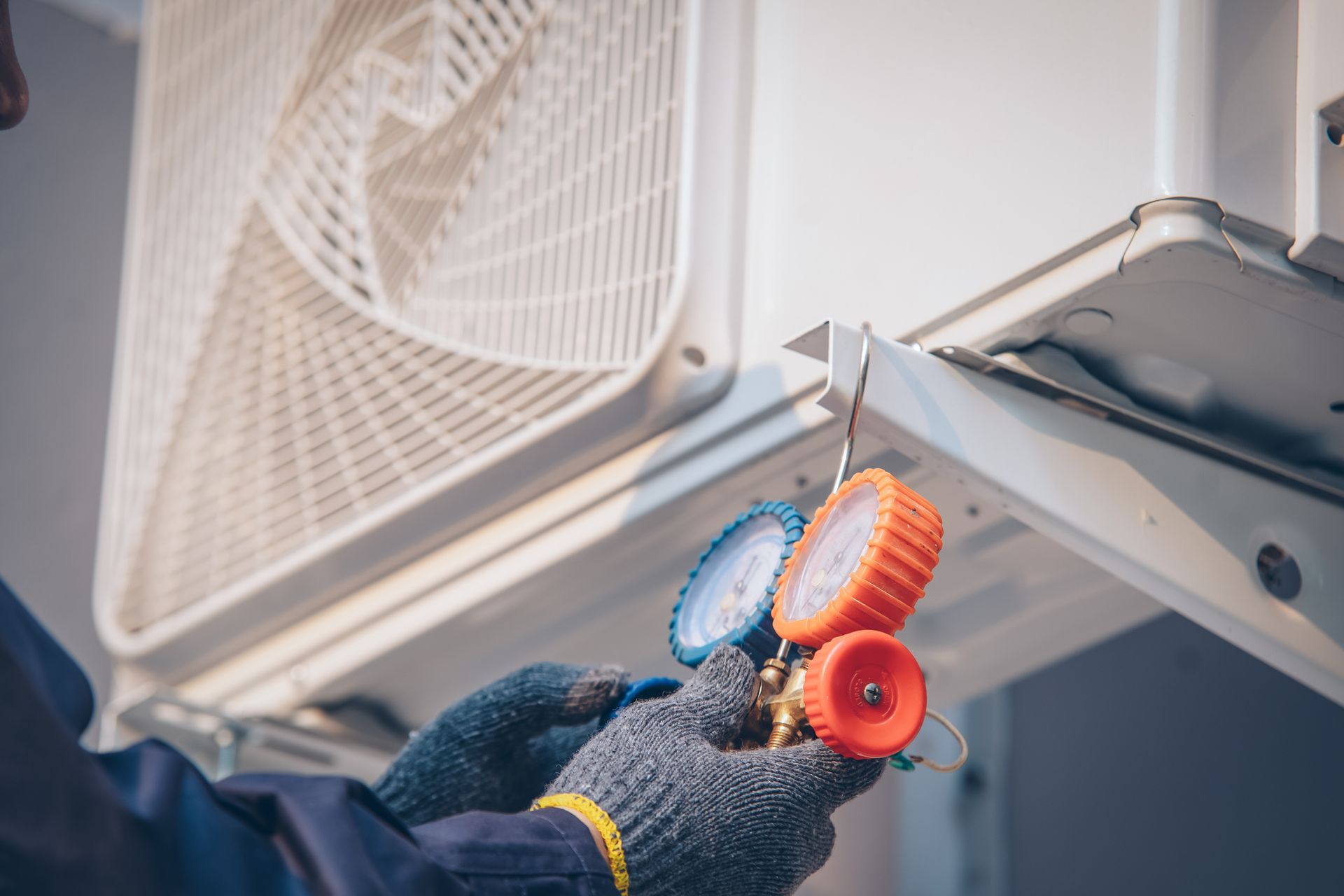 A person with gloves checks an air conditioner's pressure with a gauge in an outdoor setting.