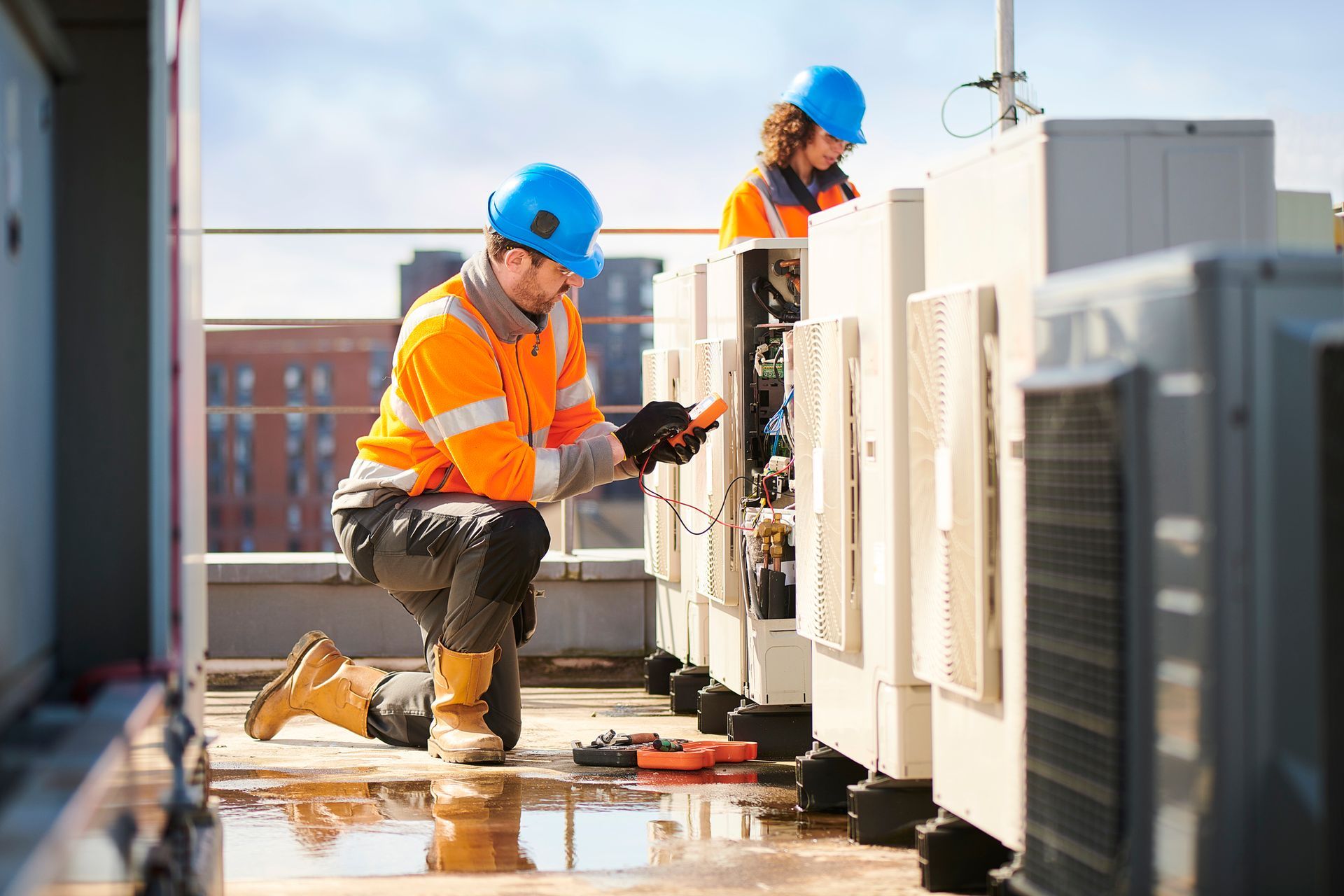 Two HVAC technicians in safety gear work on rooftop units.