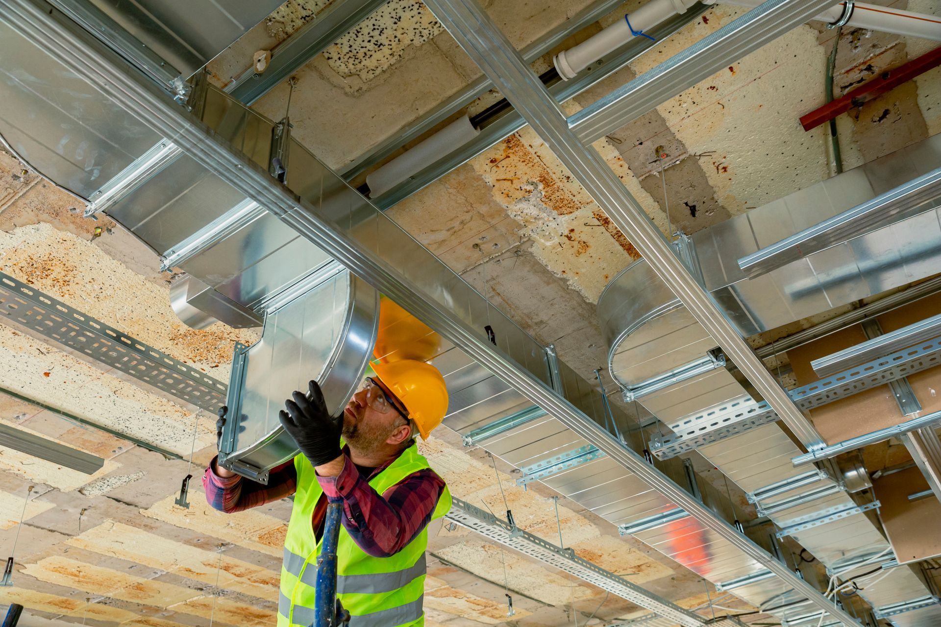 Construction worker in hard hat, vest, installing ventilation ductwork in a ceiling.