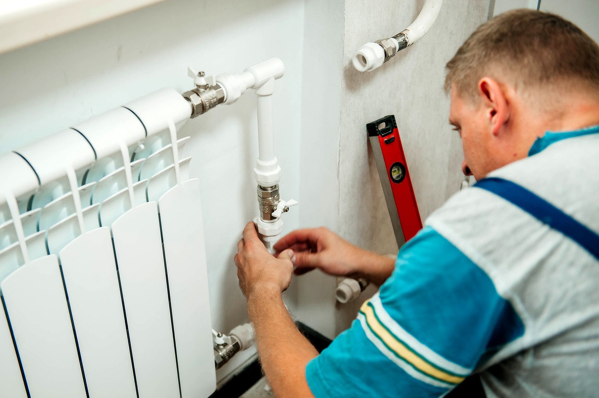 Plumber in blue shirt working on pipes near a radiator with a level on the wall.