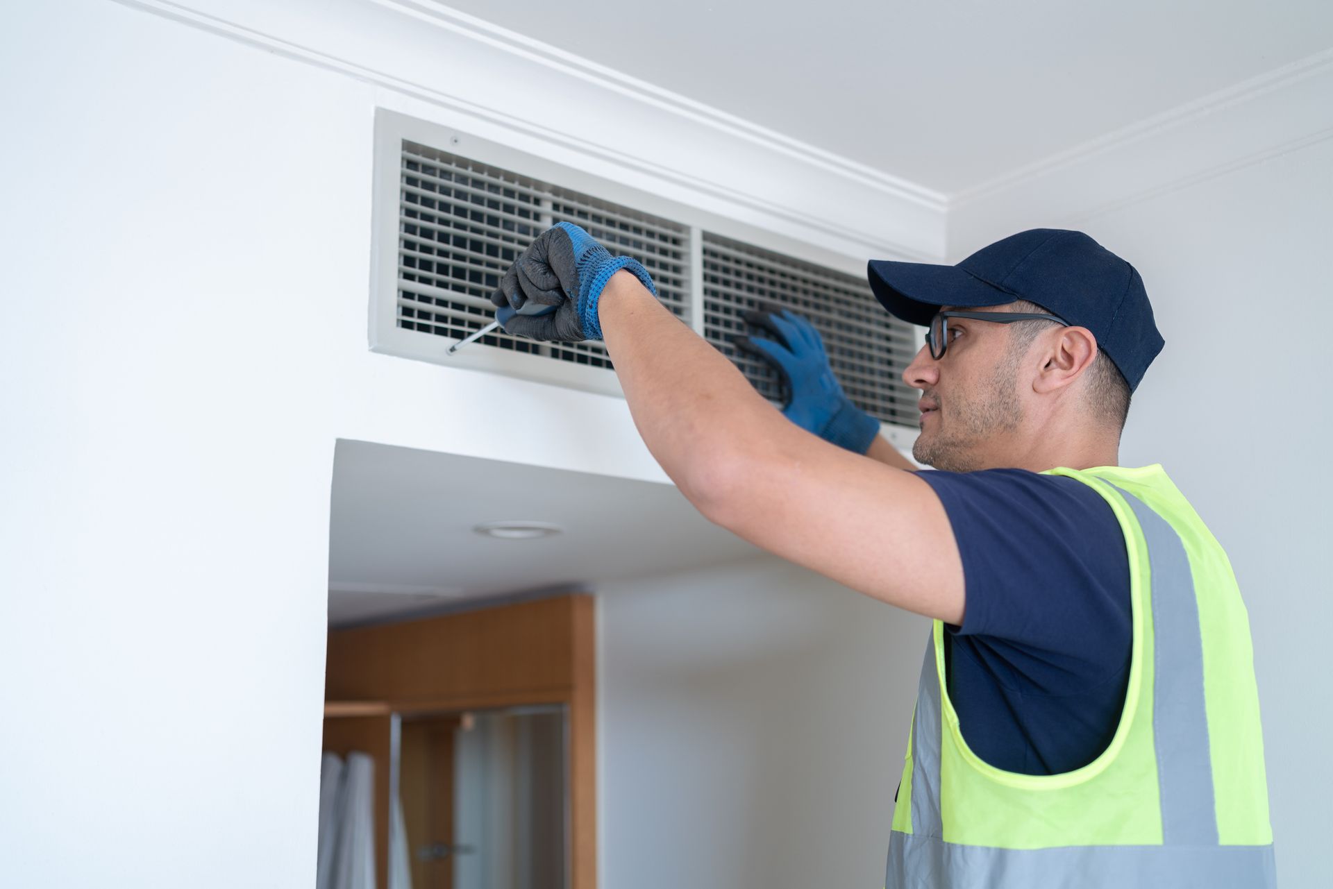 A man in safety gear cleaning an air vent. White wall, blue gloves, and a cap are visible.