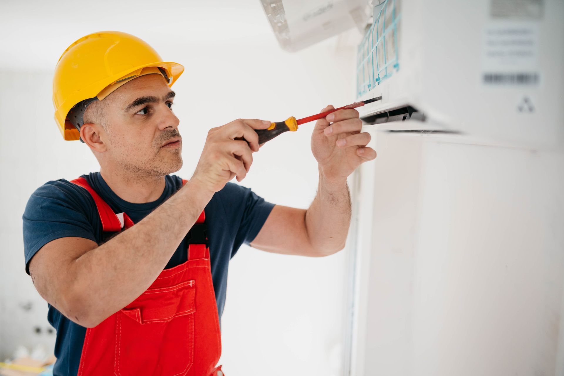 Construction worker in a yellow helmet using a screwdriver on an AC unit on a wall.