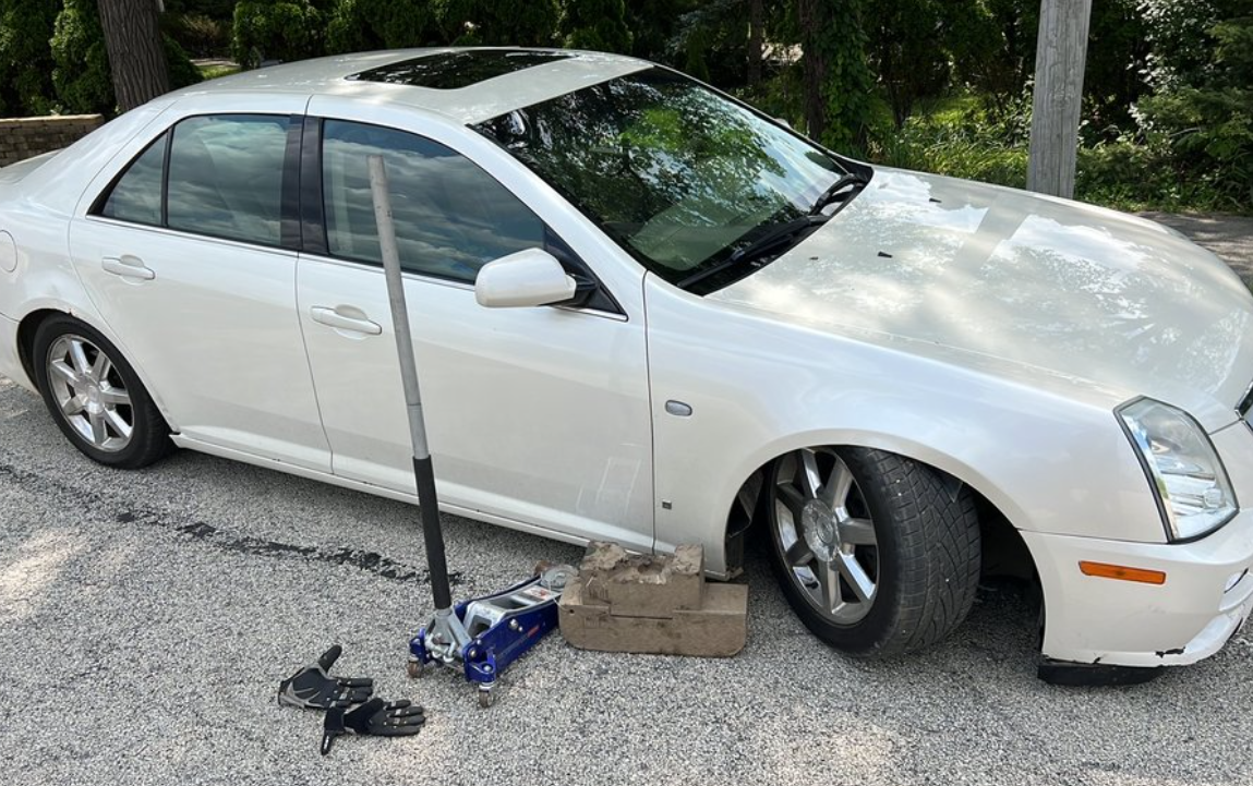 A broken down white car getting a tire change on the side of a road.