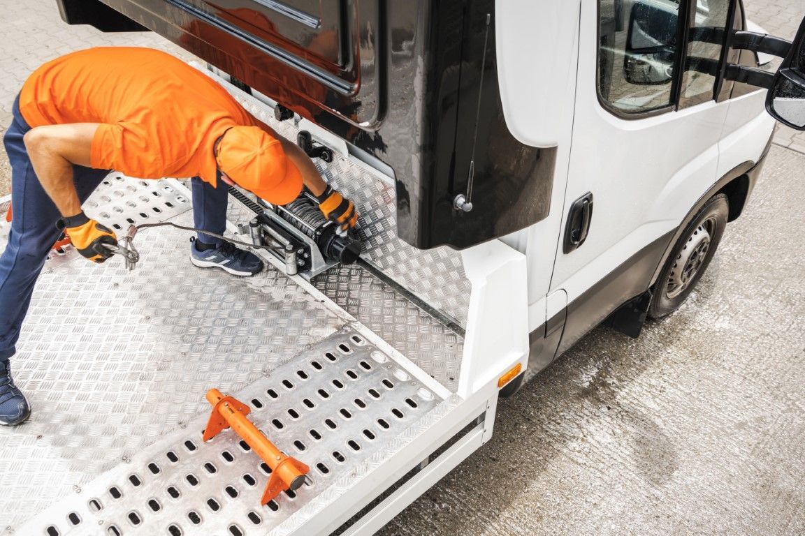 A tow truck driver in orange shirt and hat works on winch on a tow truck's ramp.