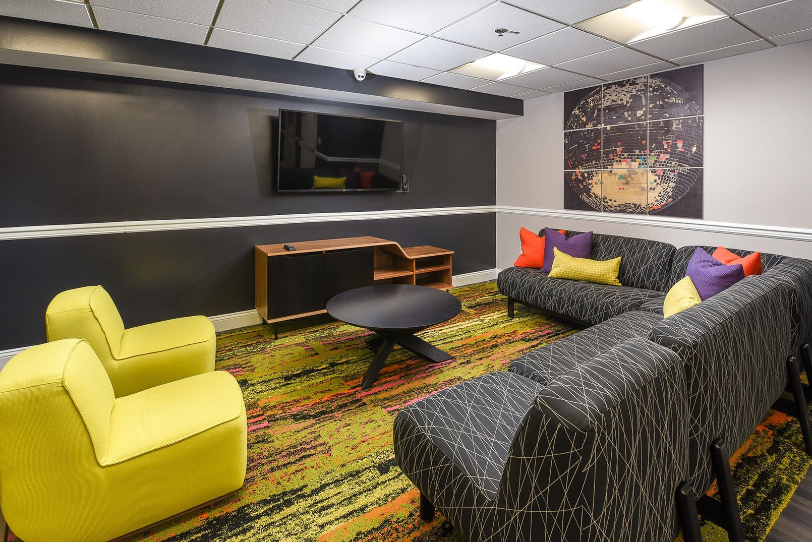 Lounge area with black and yellow patterned furniture, dark gray wall with TV, and colorful rug at Glenmont Station in Silver Spring, MD.