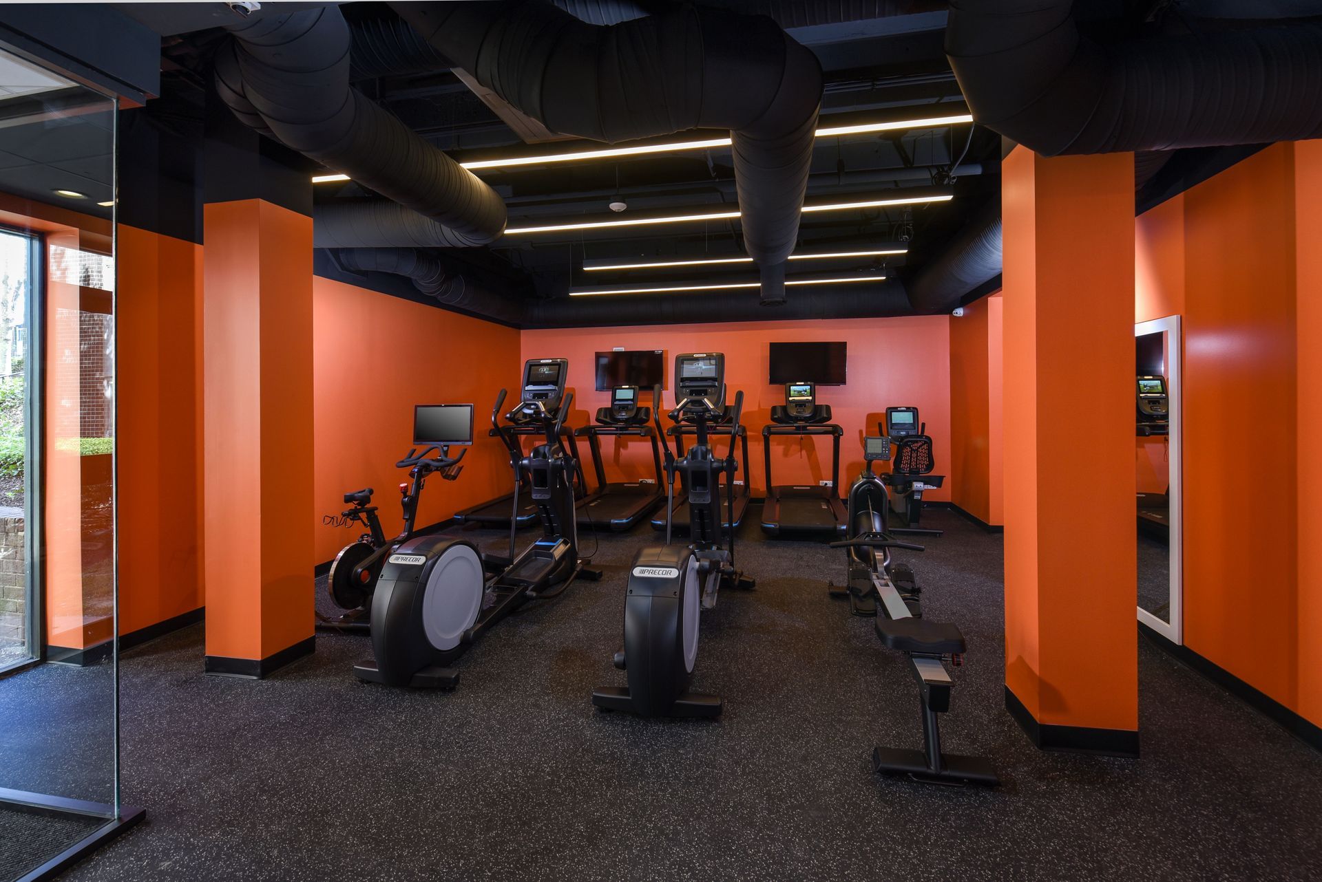 Orange-walled gym with black ceiling, featuring elliptical machines, treadmills, rowing machine, and TVs at Glenmont Station in Silver Spring, MD.
