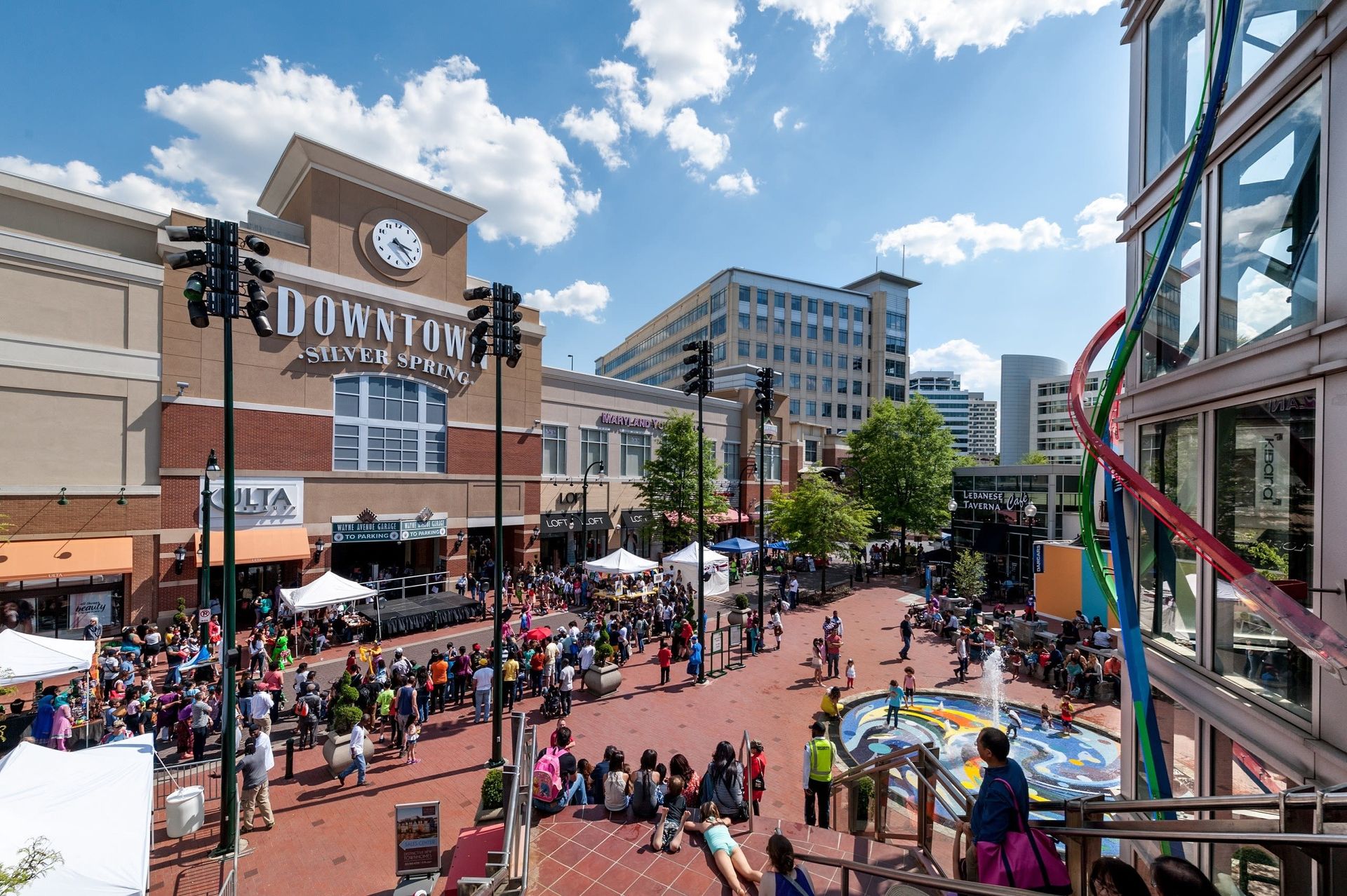 Downtown plaza with event, brick ground, crowd, buildings, fountain, sunny sky at Glenmont Station in Silver Spring, MD.