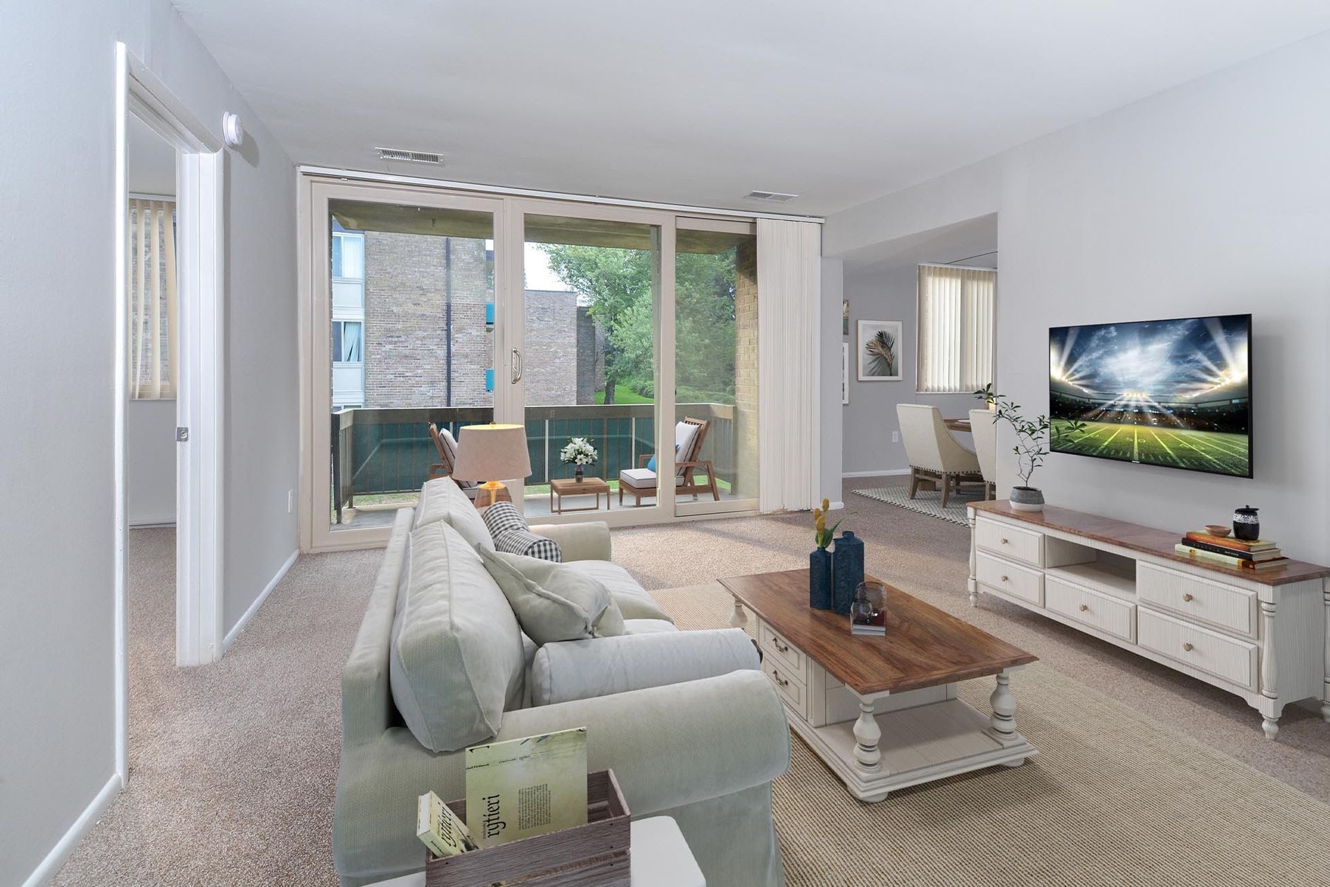 Living room with sofa, TV, coffee table, and balcony access. Beige and white tones at Glenmont Station in Silver Spring, MD.