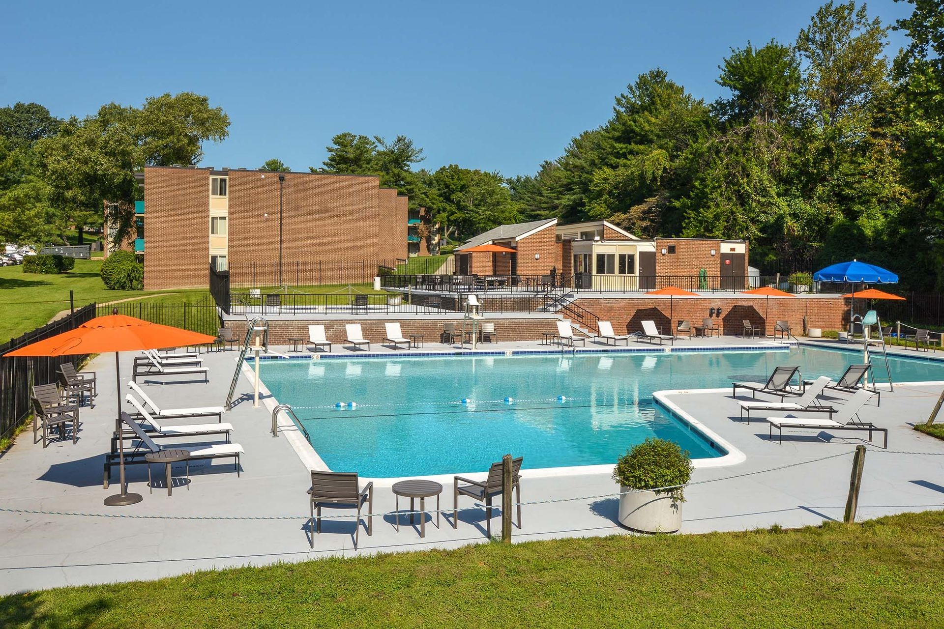 Swimming pool with lounge chairs, orange and blue umbrellas, and a brick building on a sunny day at Glenmont Station in Silver Spring, MD.
