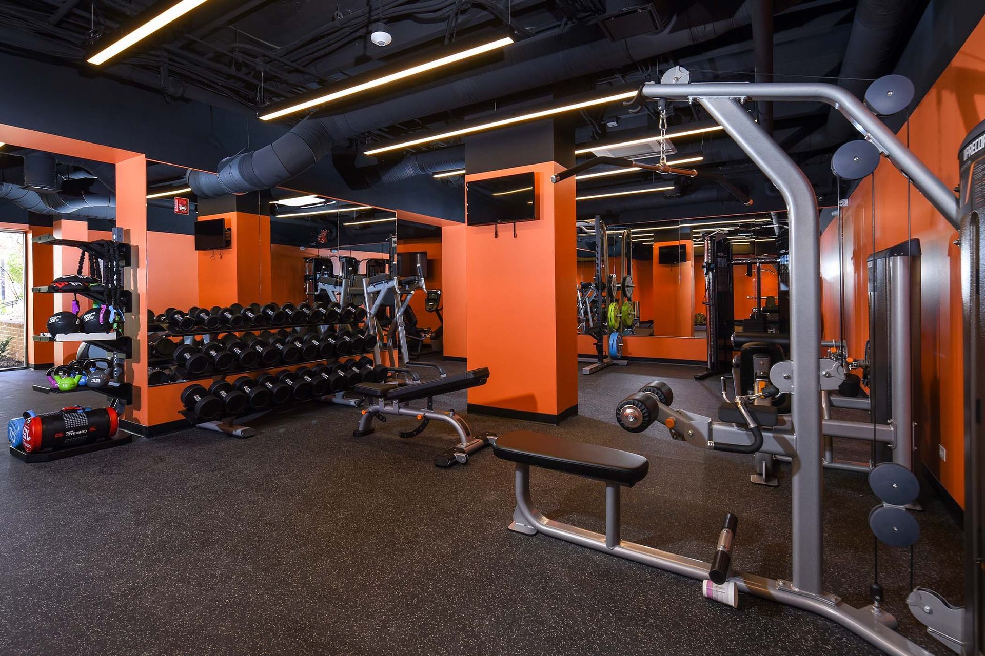 Gym interior with weight machines, free weights, and black rubber flooring, accented with orange walls at Glenmont Station in Silver Spring, MD.