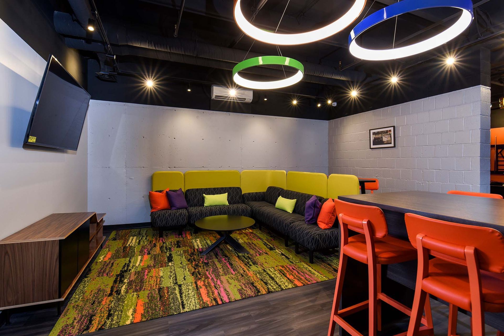Lounge area with a colorful sectional, high-top table, and vibrant rug. Overhead, colorful ring lights at Glenmont Station in Silver Spring, MD.