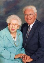 Elderly couple, woman in turquoise jacket, man in blue suit, smiling, holding hands.