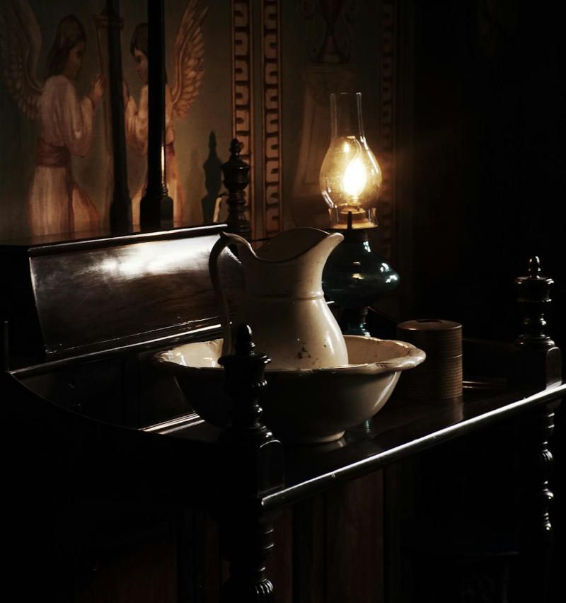 A lit oil lamp casts light on a wash basin and pitcher on a dark wooden cabinet, against a mural backdrop.