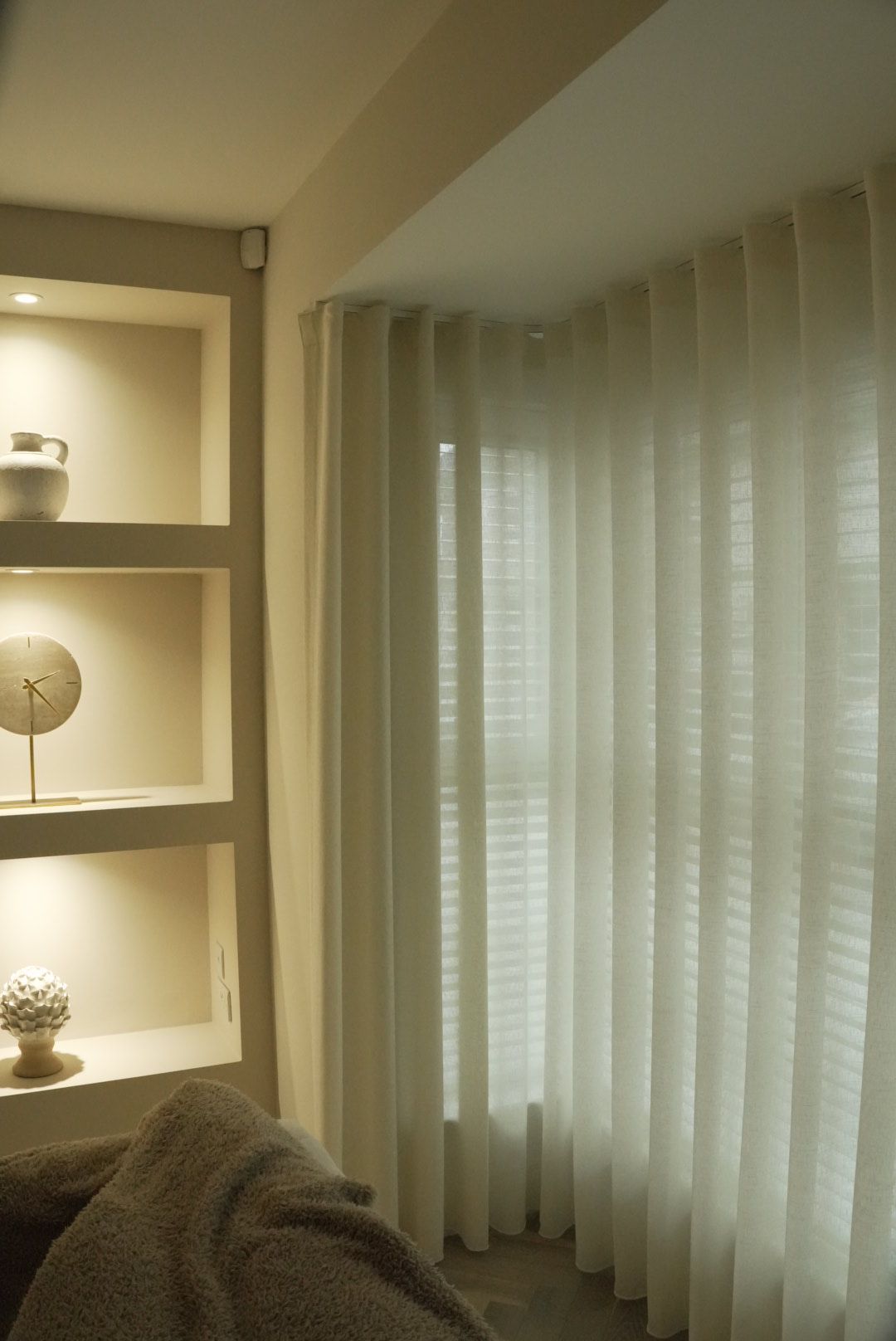 White curtains next to a window, lit shelves with decor on the left, and part of a couch.