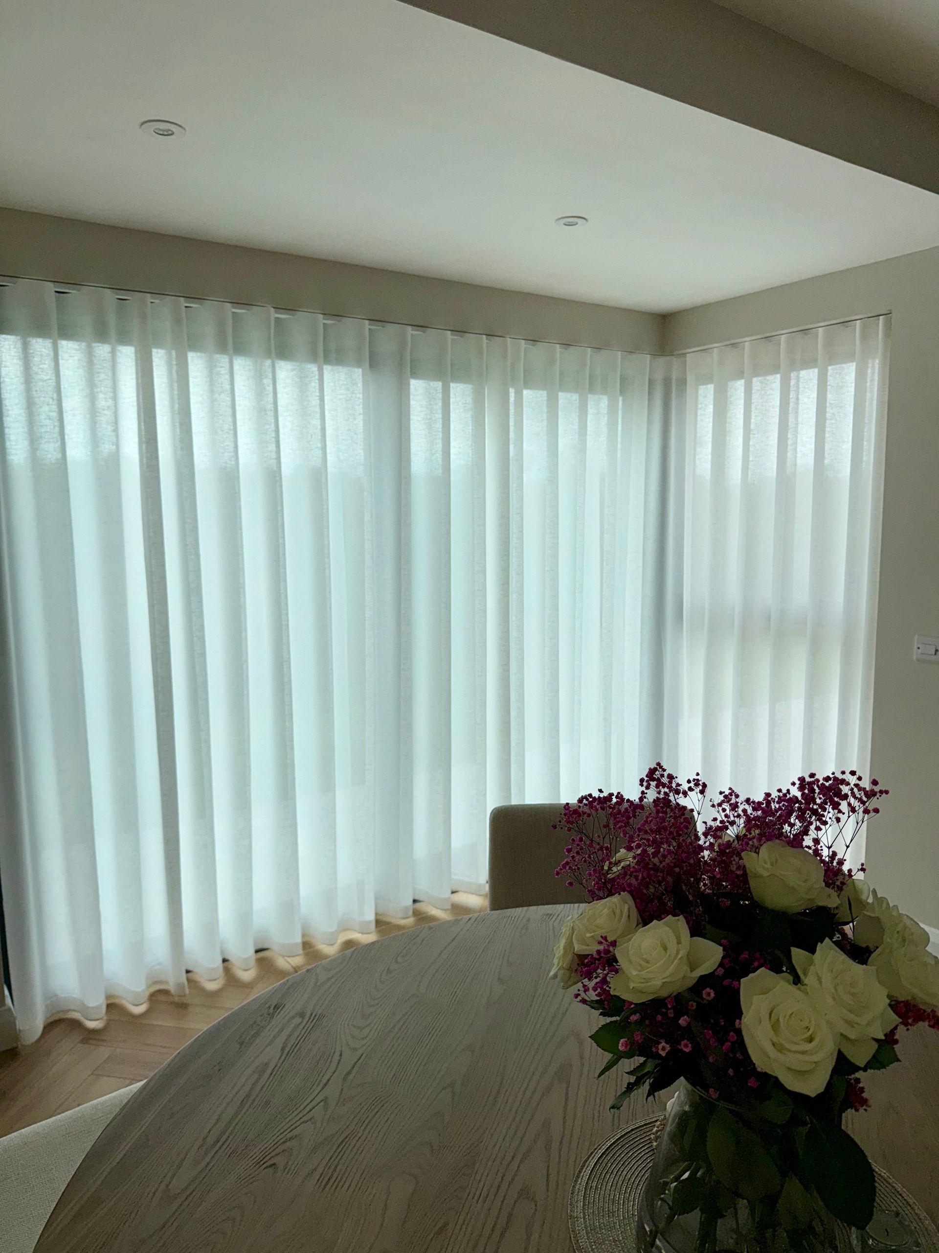 White sheer curtains in front of windows; a table with a bouquet of pink and white flowers.
