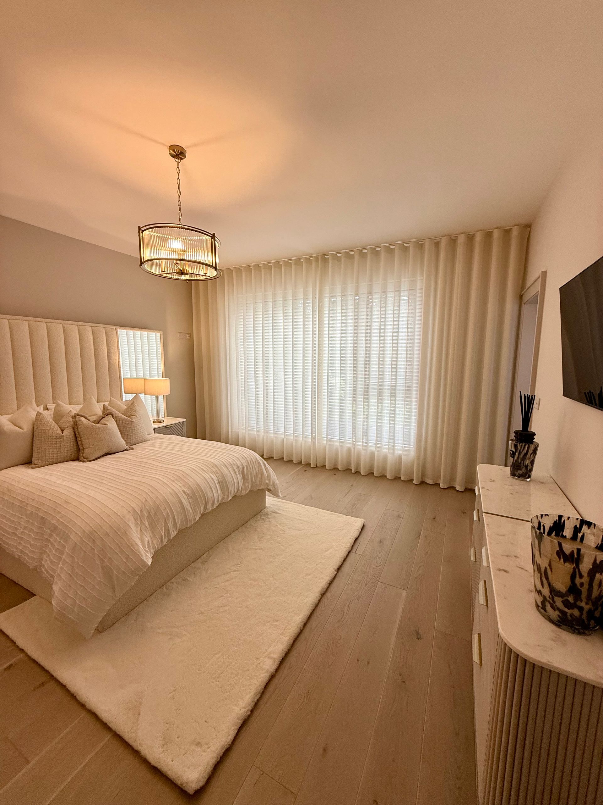 Elegant bedroom with white bed, rug, curtains, and dresser. Gold chandelier, light wood floors.