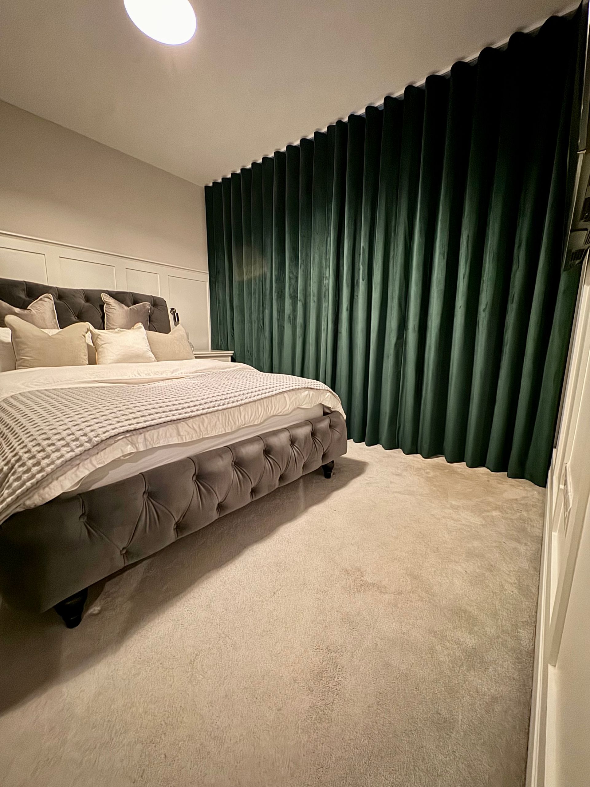 Bedroom with a gray button-tufted bed, green velvet curtains, and light gray carpet.