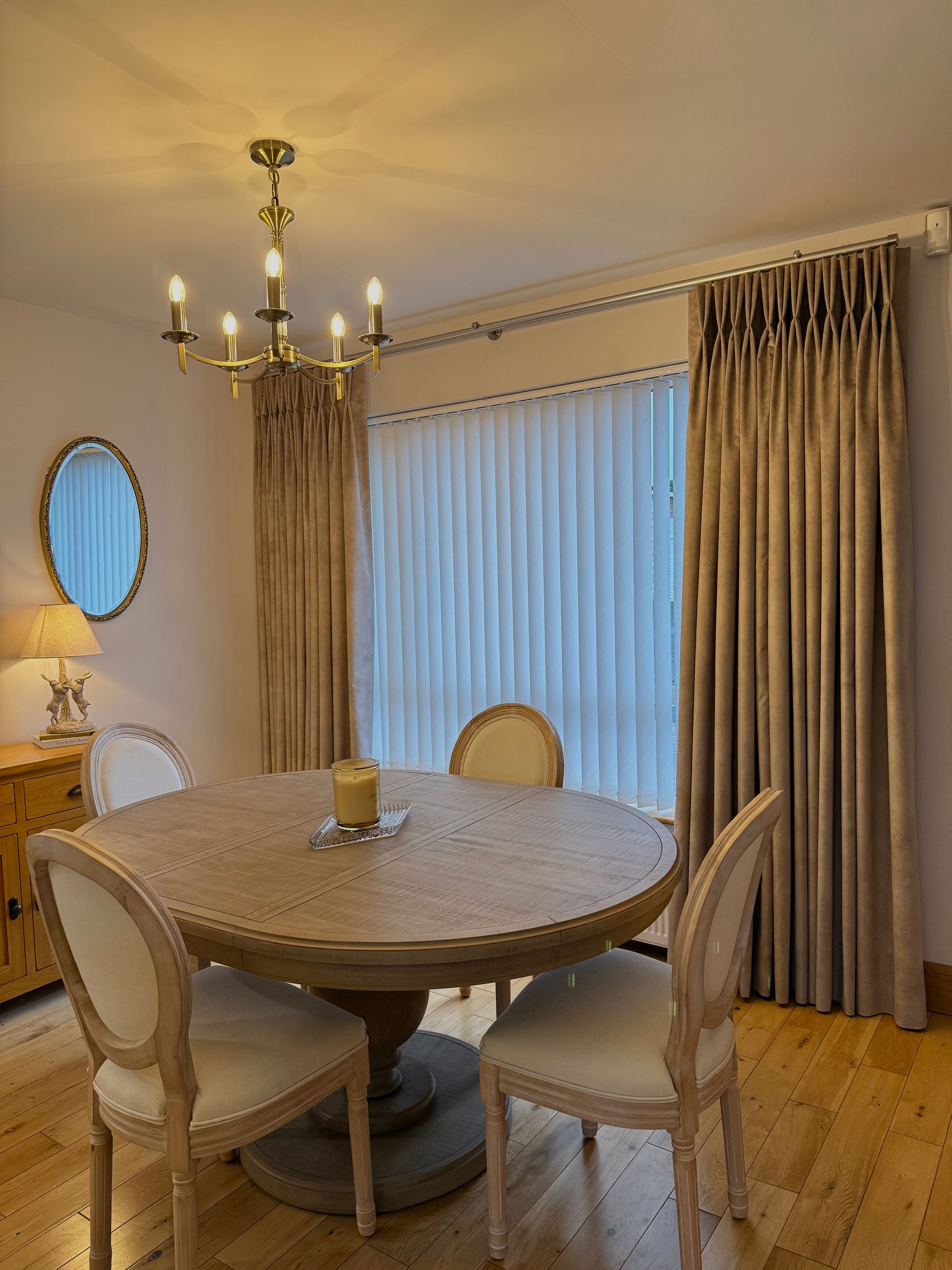 Dining room with round table, chairs, and gold chandelier. Beige curtains frame a window.