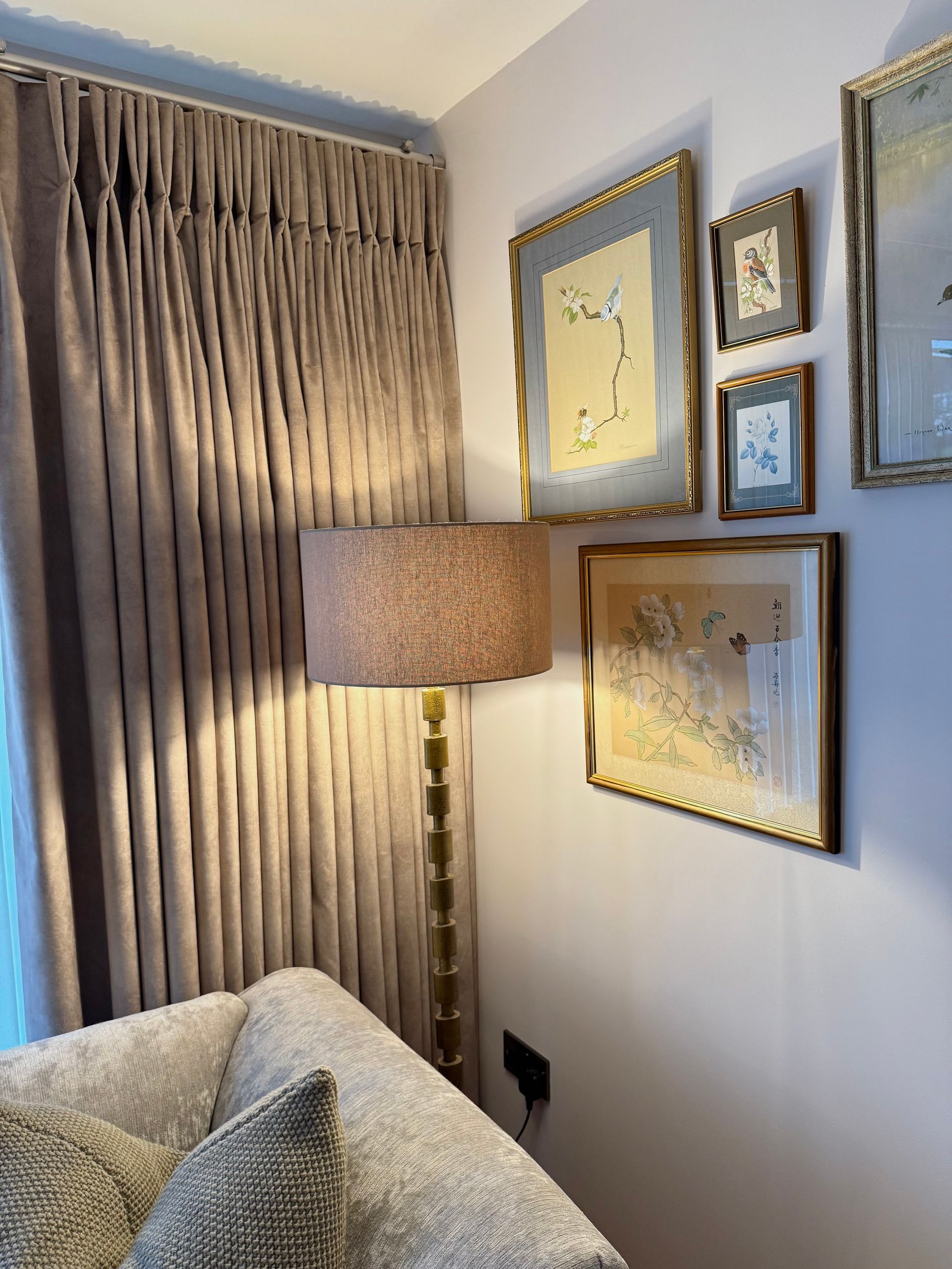 Corner with a floor lamp and framed art on a white wall. Beige curtains on the left, a sofa in the bottom left.