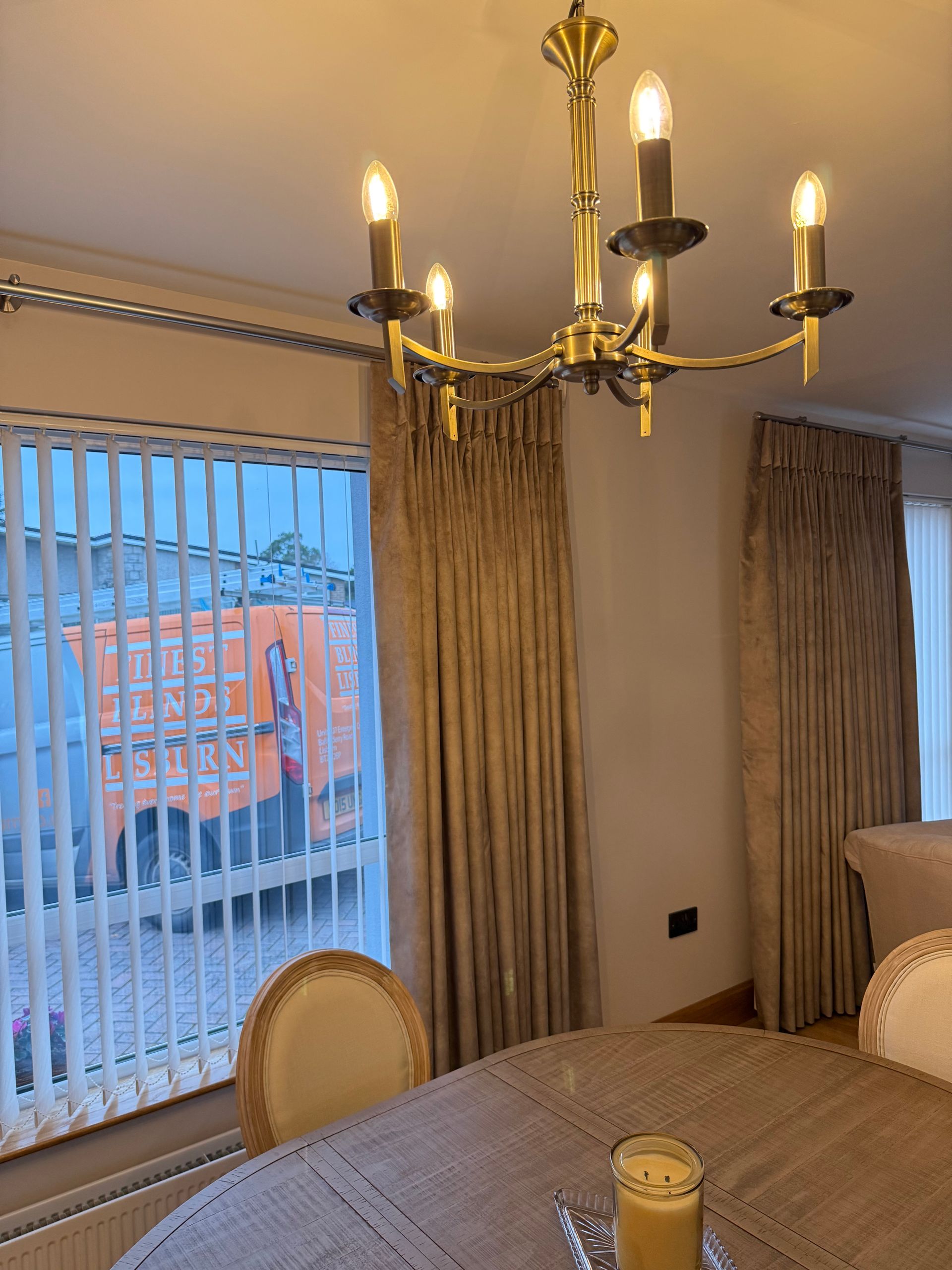 Dining room with chandelier, curtains, and table. Vertical blinds and an orange truck visible through the window.