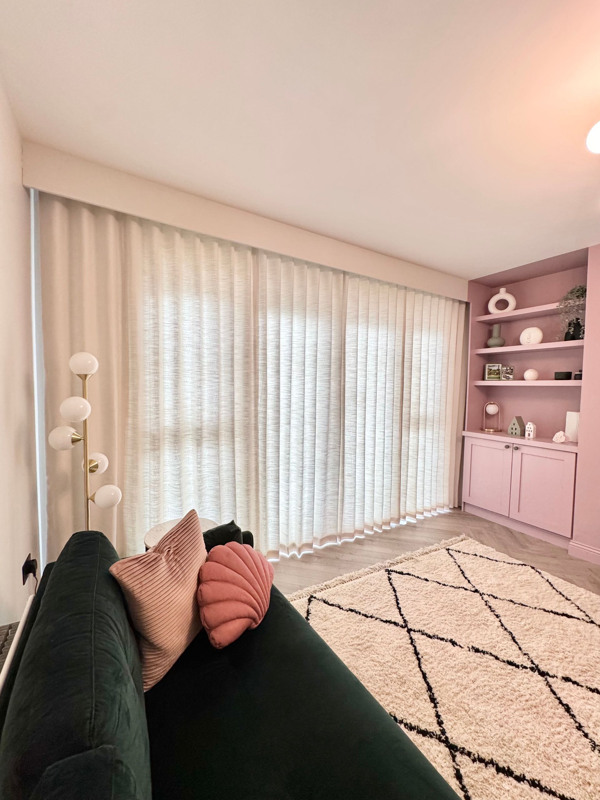 Living room with white curtains, pink bookcase, and dark green sofa with pink pillows.