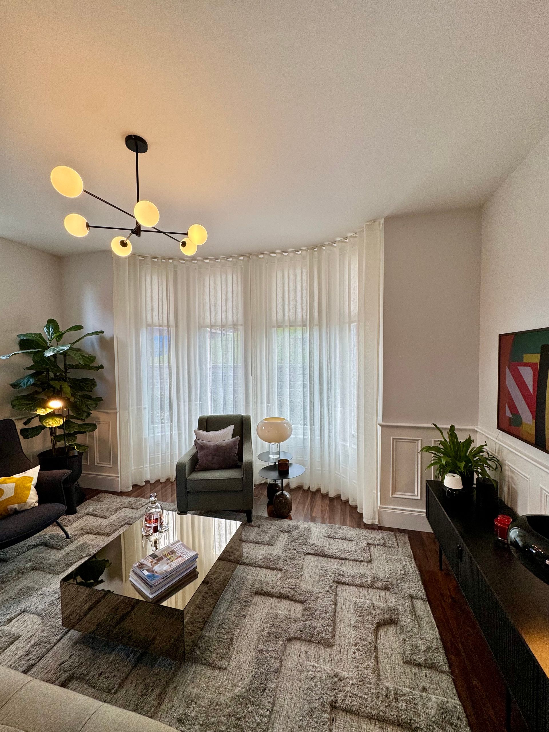 Living room with modern decor: gray rug, curved sheer curtains, chandelier, and a chair.
