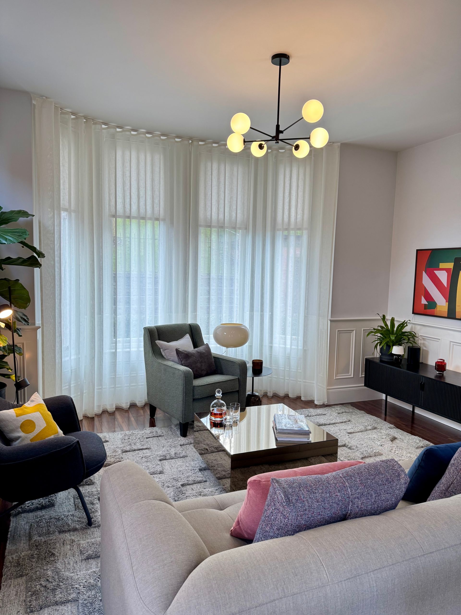 Living room with sofa, chairs, rug, and modern light fixture. Large sheer curtains cover the windows.