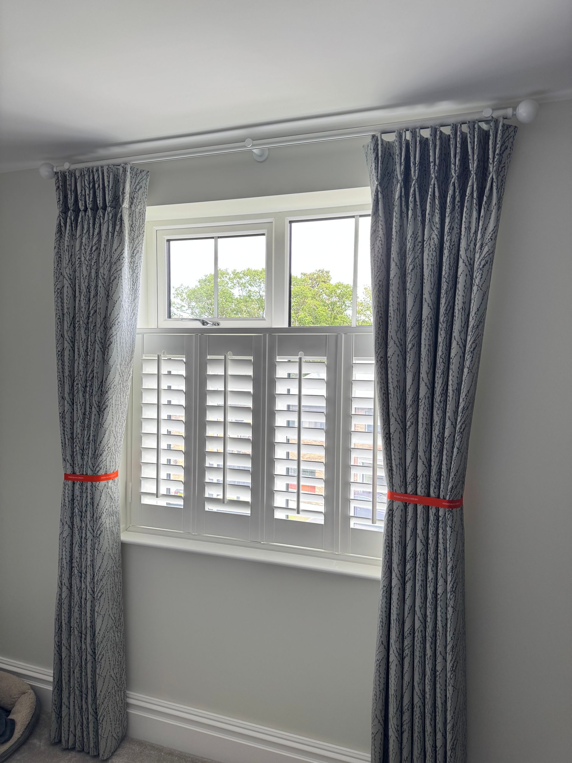 Window with white shutters and patterned curtains, held by orange ties.