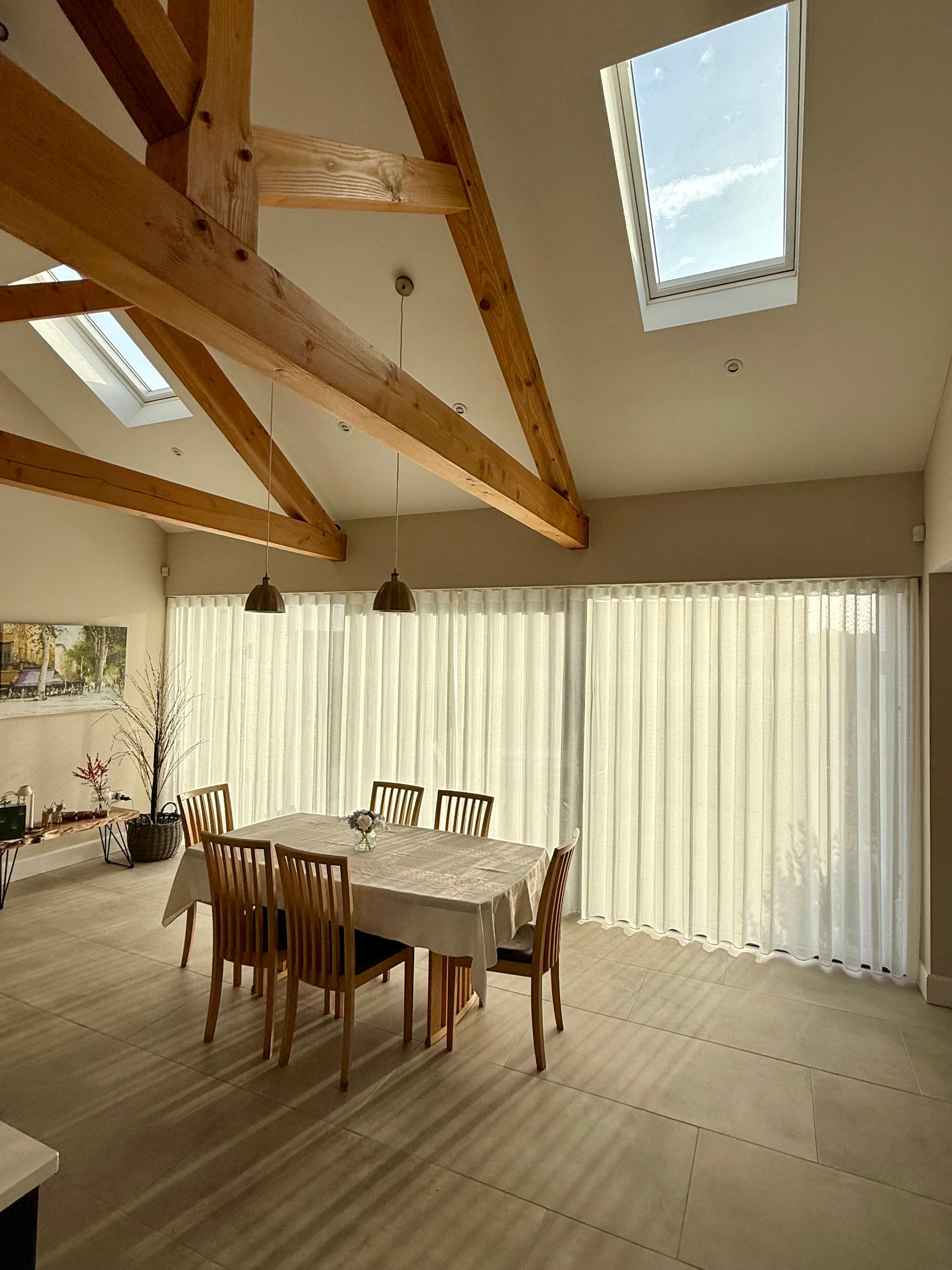 Dining room with light-colored curtains, a wooden table with chairs, skylights, and exposed wooden beams.