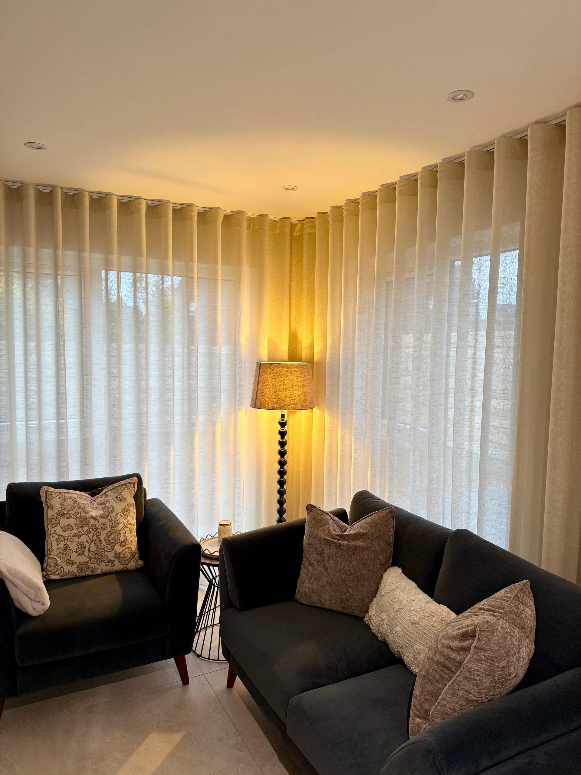 Living room with cream-colored curtains framing a black sofa and armchair, a lamp, and decorative pillows.