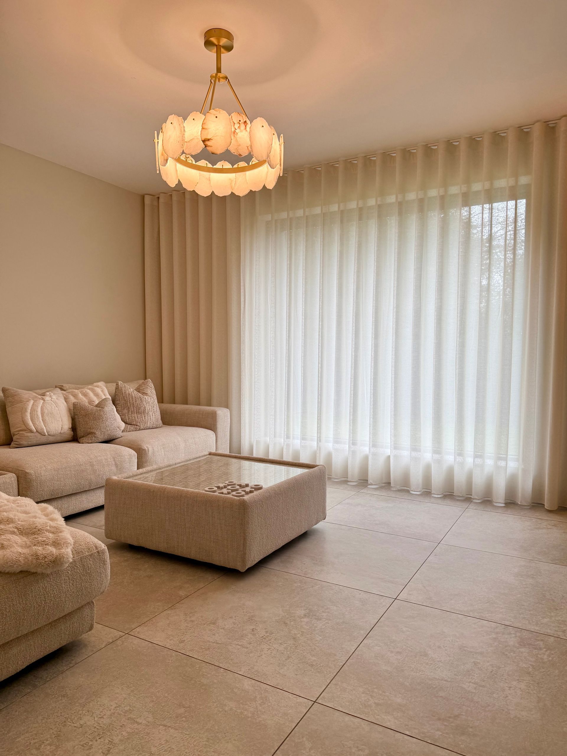 Living room with beige furniture, curtains, and chandelier, neutral color palette.