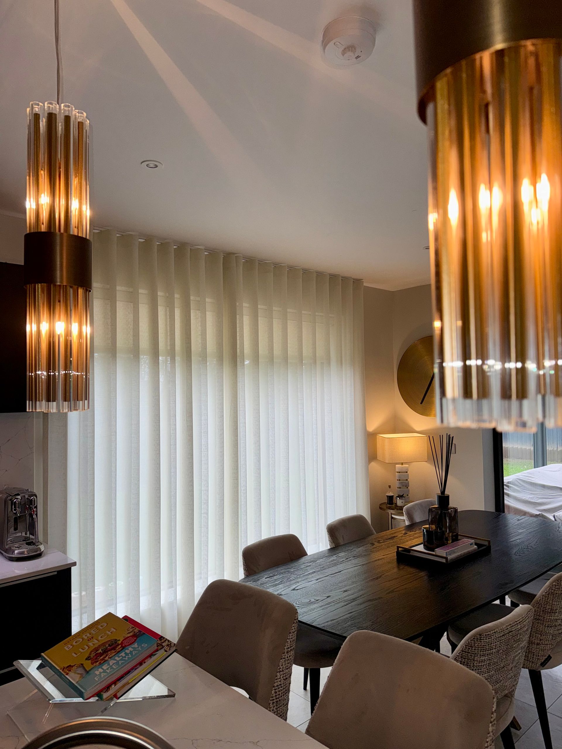 Dining room with a dark table, velvet chairs, sheer curtains, and gold pendant lights.