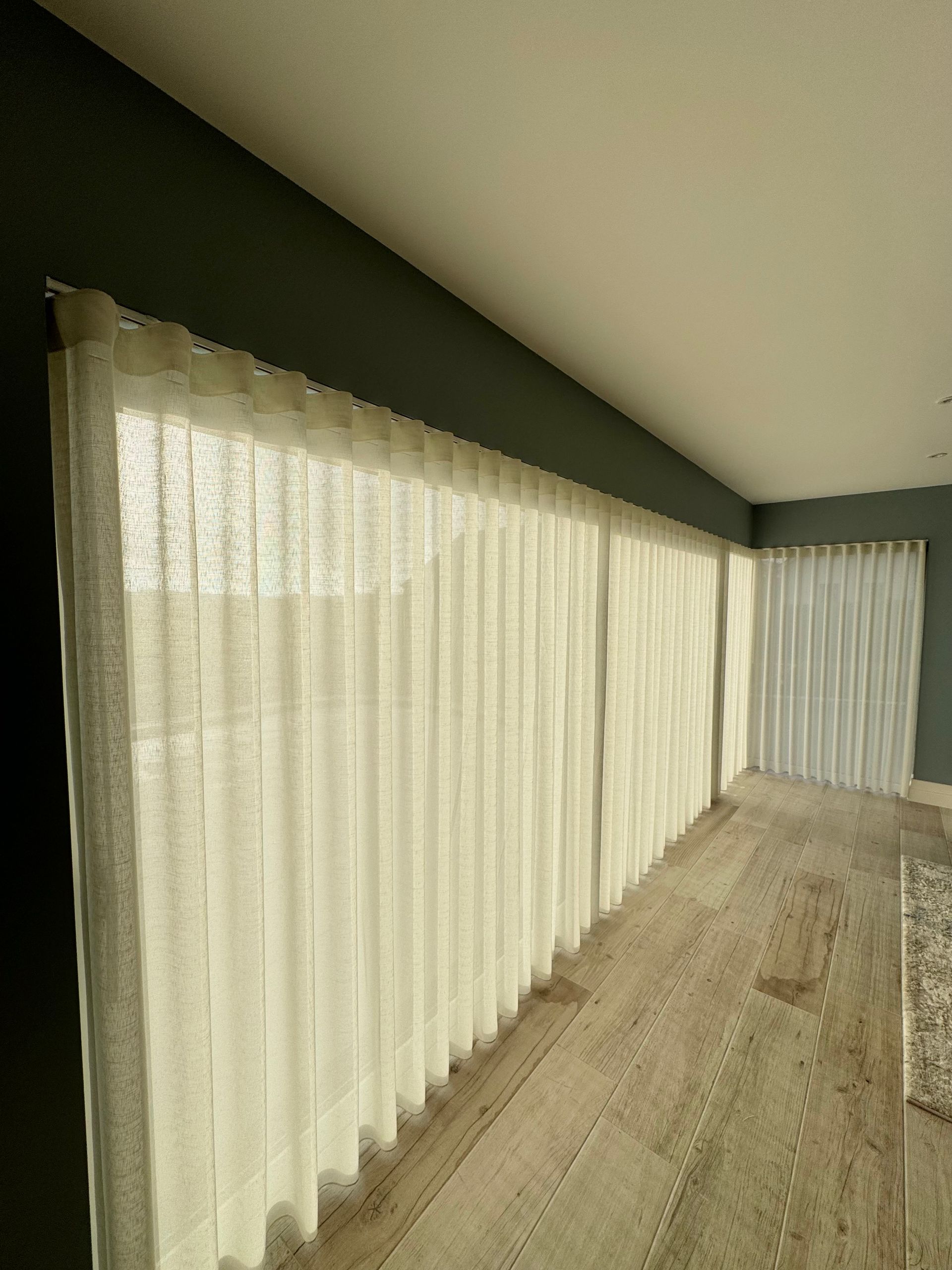 Cream-colored sheer curtains cover a long window on a dark green wall, alongside a light-colored wood floor.