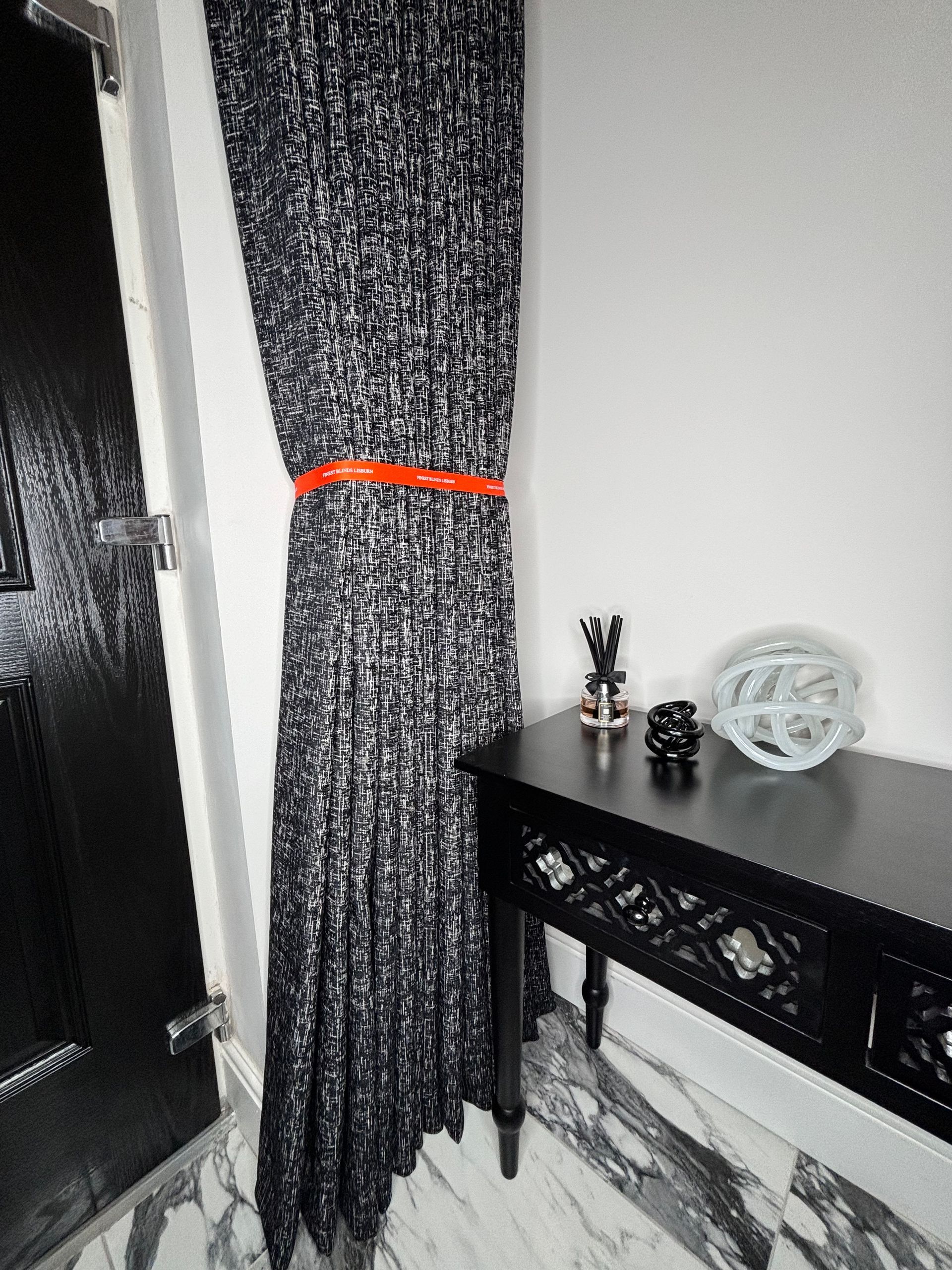Black and white patterned curtain held with an orange band, next to a black table with decor.