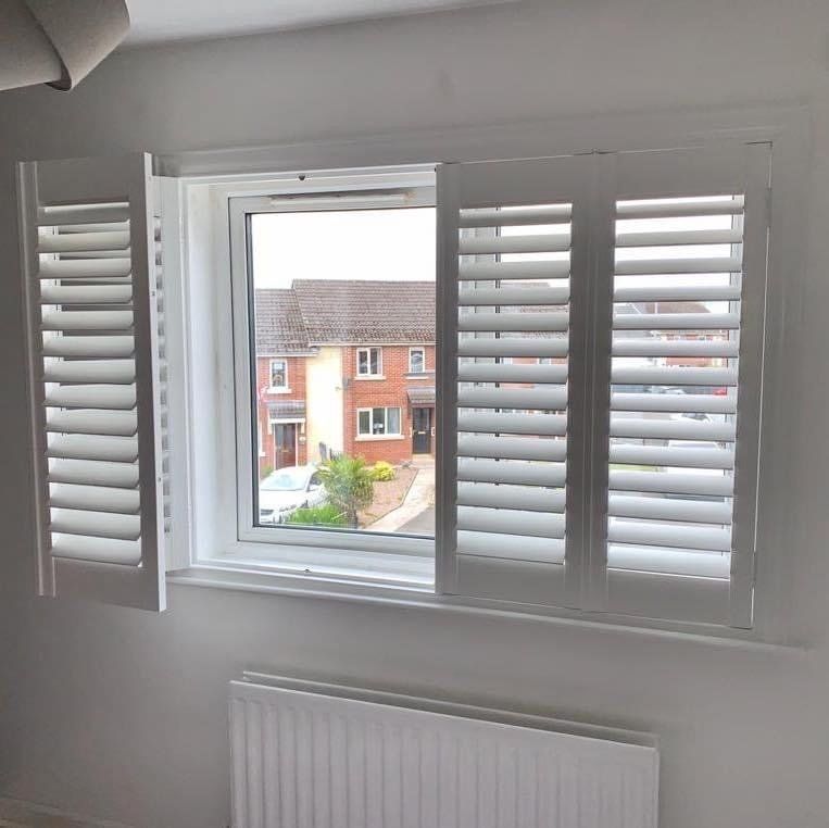 A bedroom window with white shutters and a view of a house.