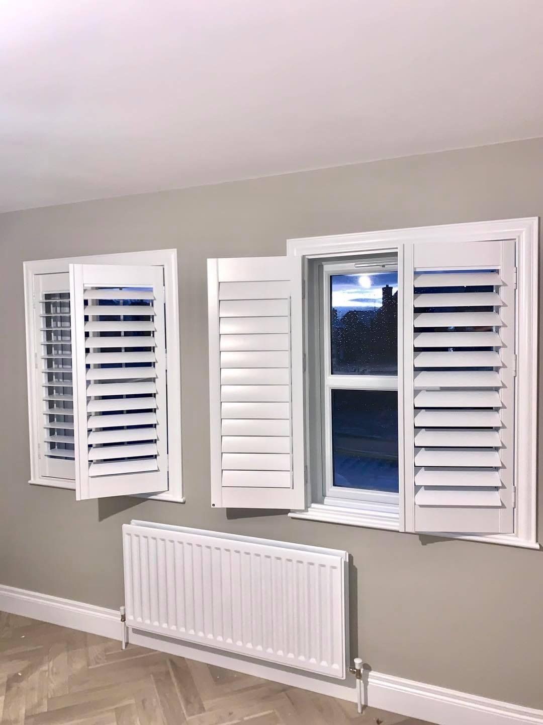 A room with two windows and a radiator with white shutters on them.