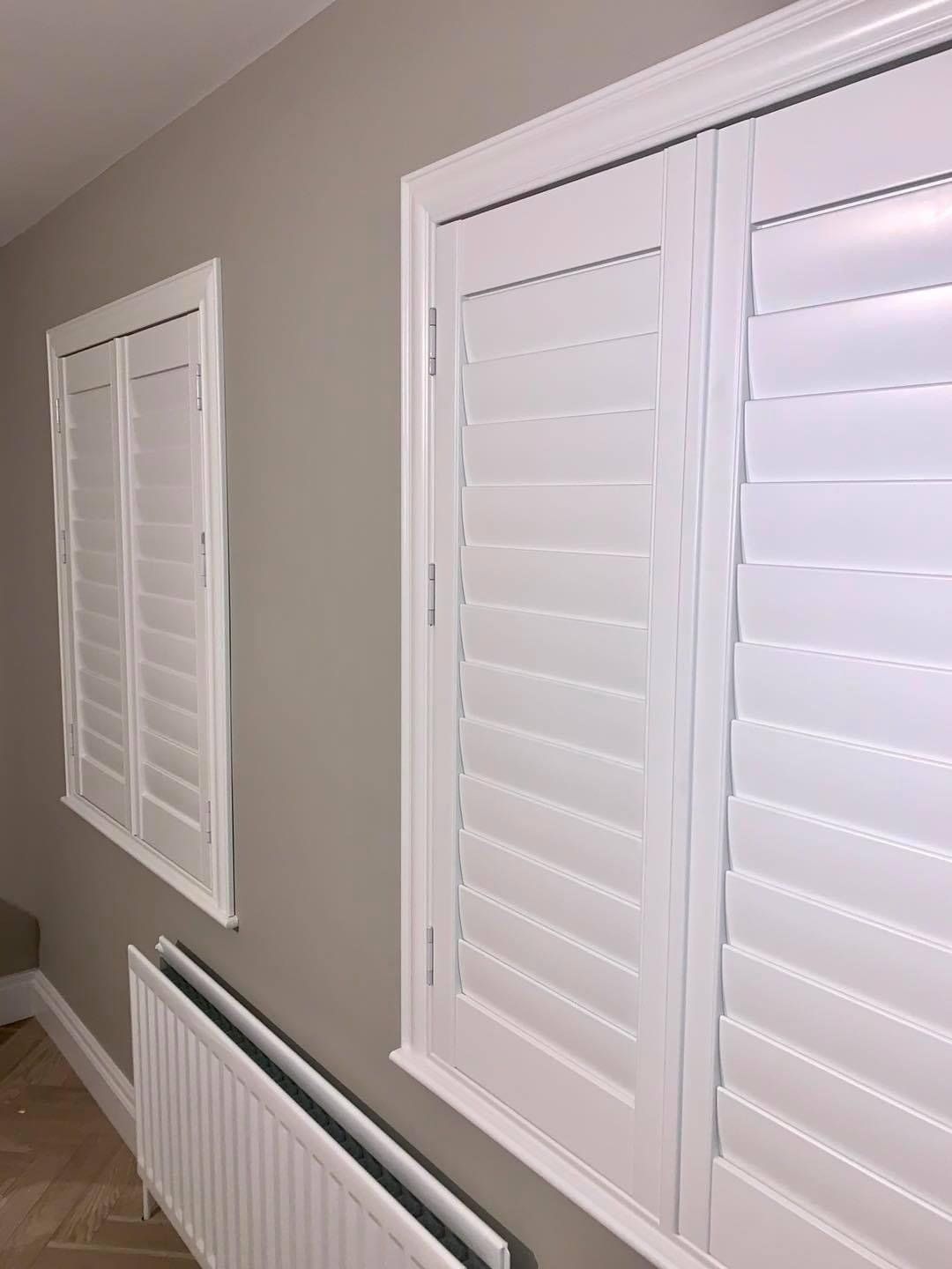 A room with two windows and a radiator with white shutters on them.