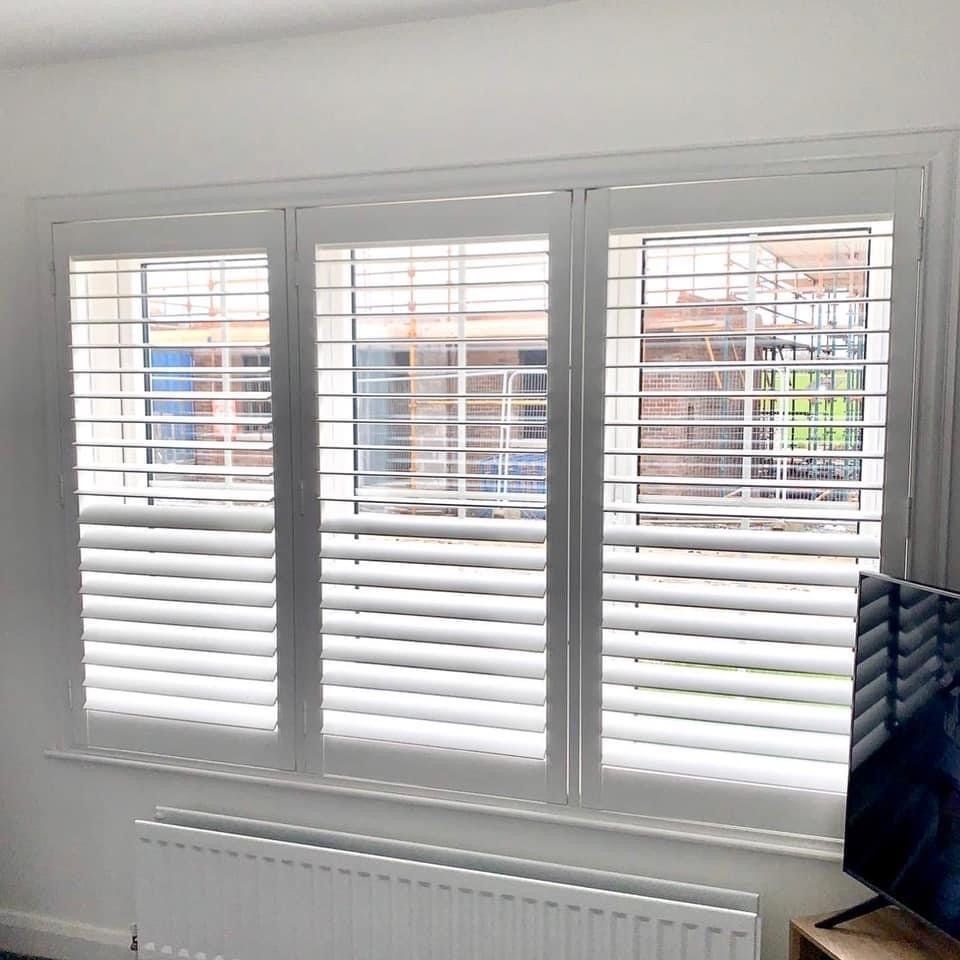 A living room with white shutters on the windows and a television.