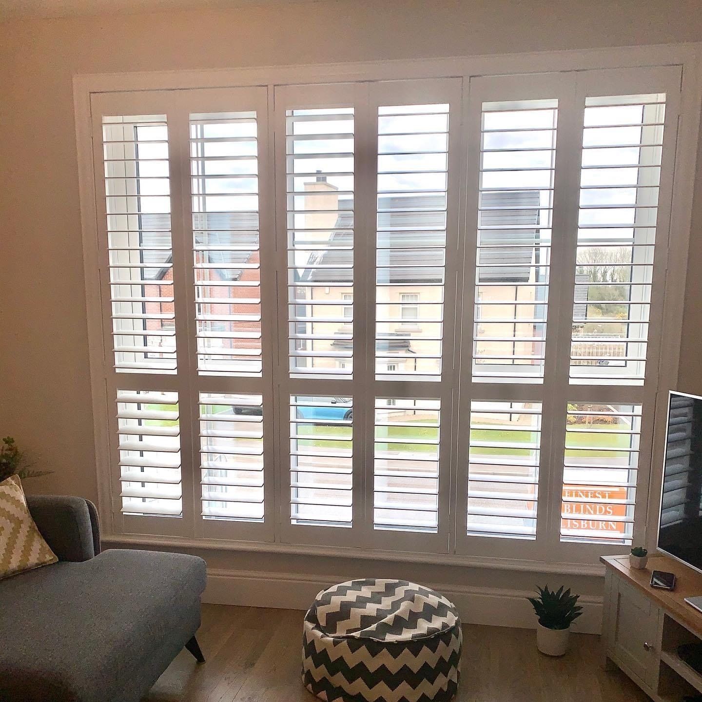 A living room with a couch , ottoman , television and shutters on the windows.