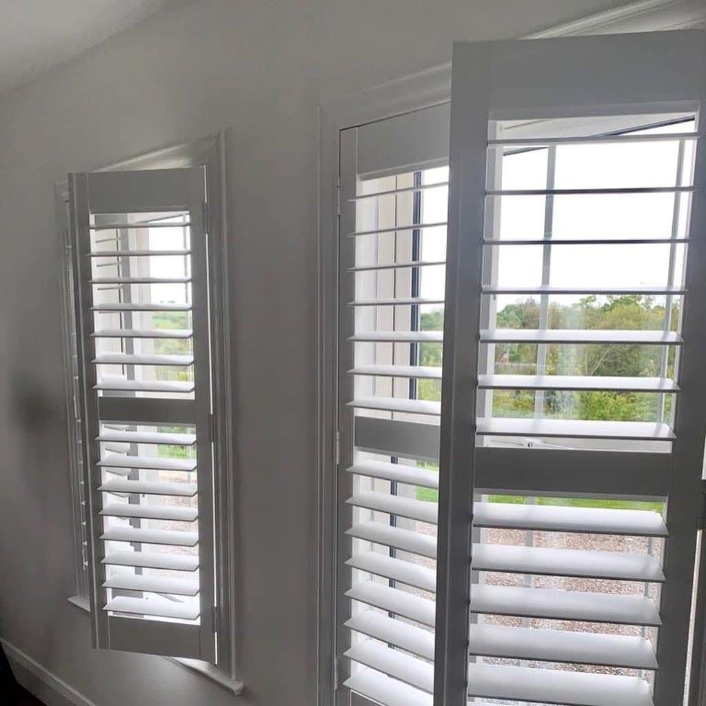 A room with two windows with white shutters on them.