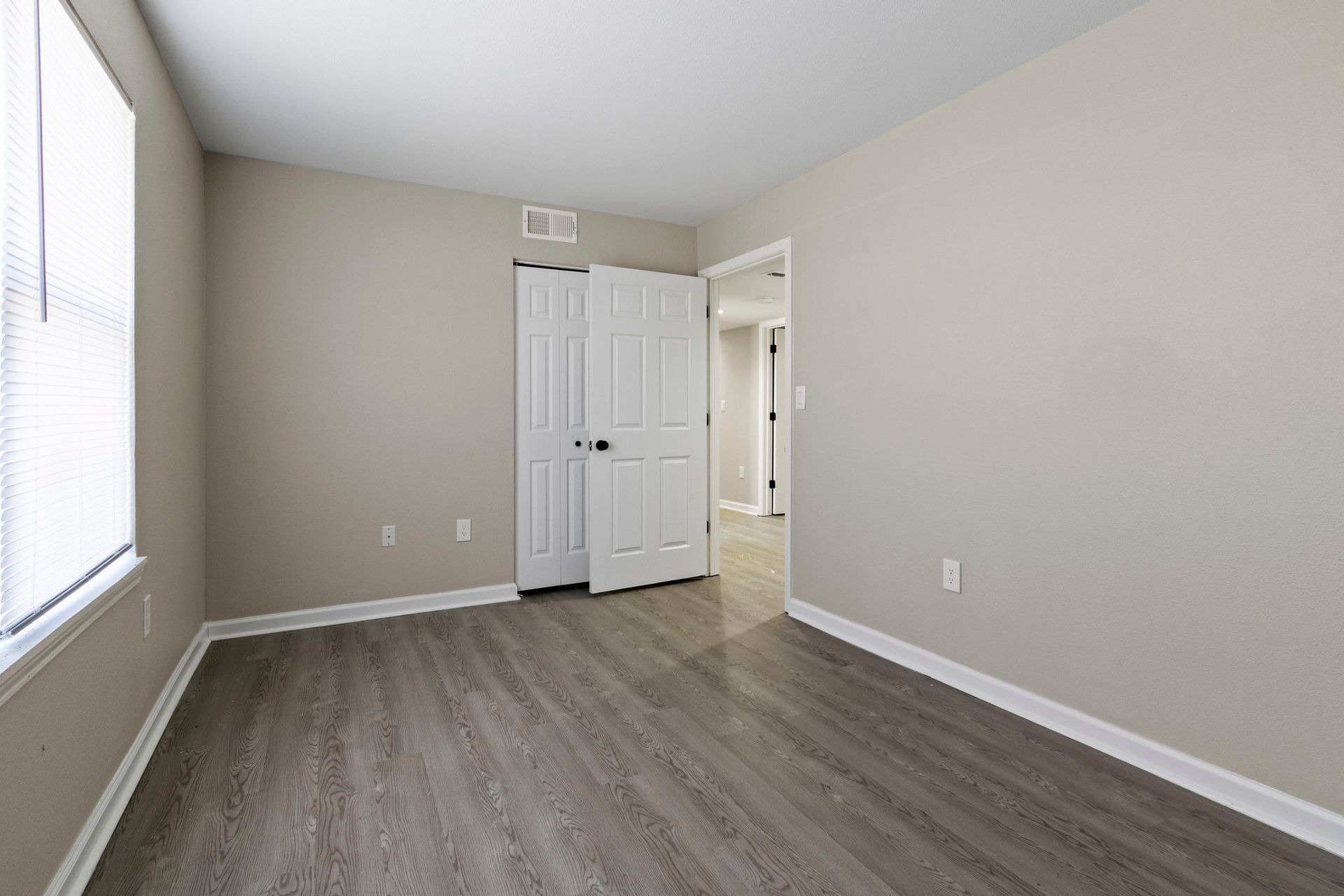 Empty bedroom with gray flooring, beige walls, white doors, and a window.