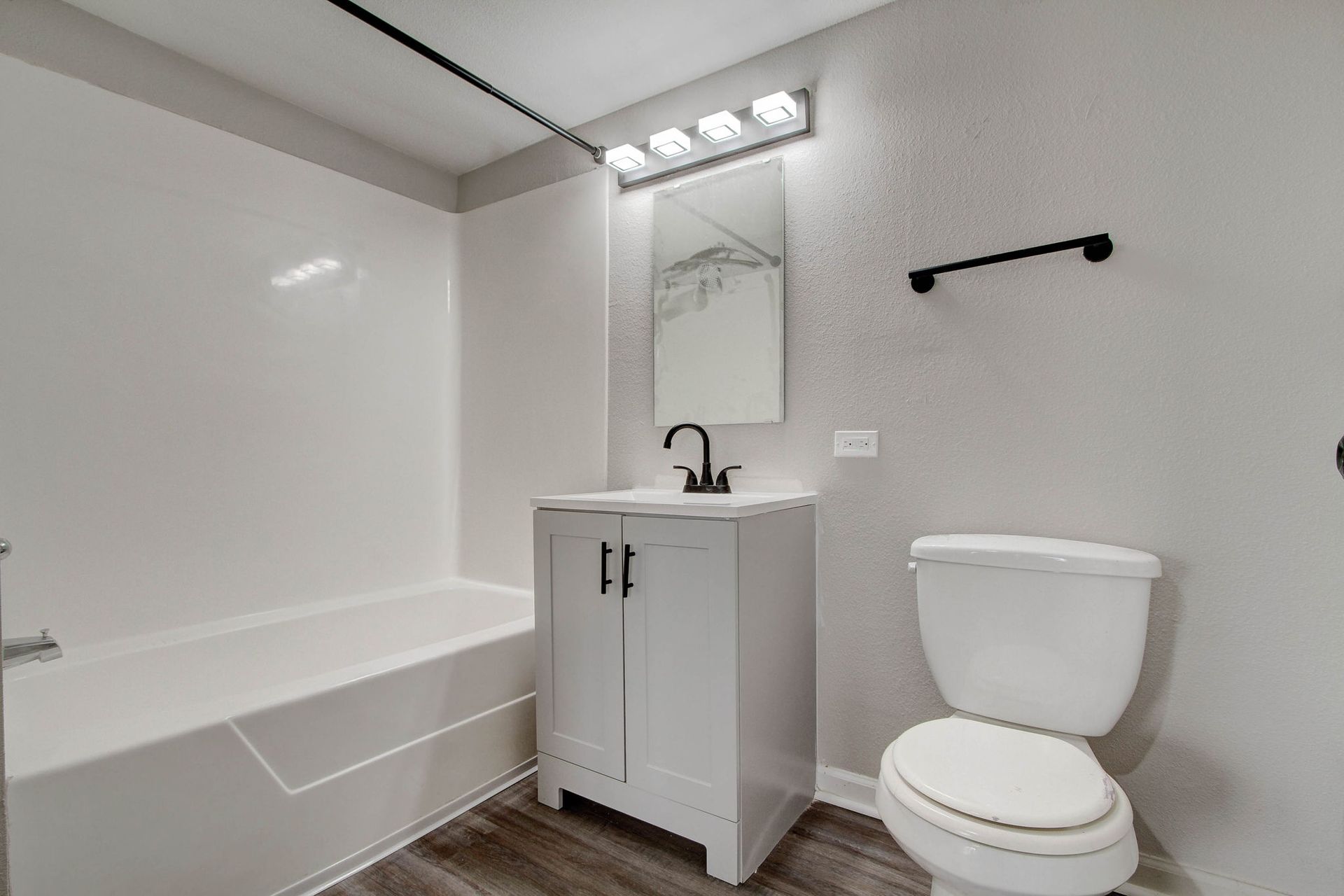Bathroom with white tub and vanity, toilet, dark fixtures, gray walls, and dark wood-look flooring.