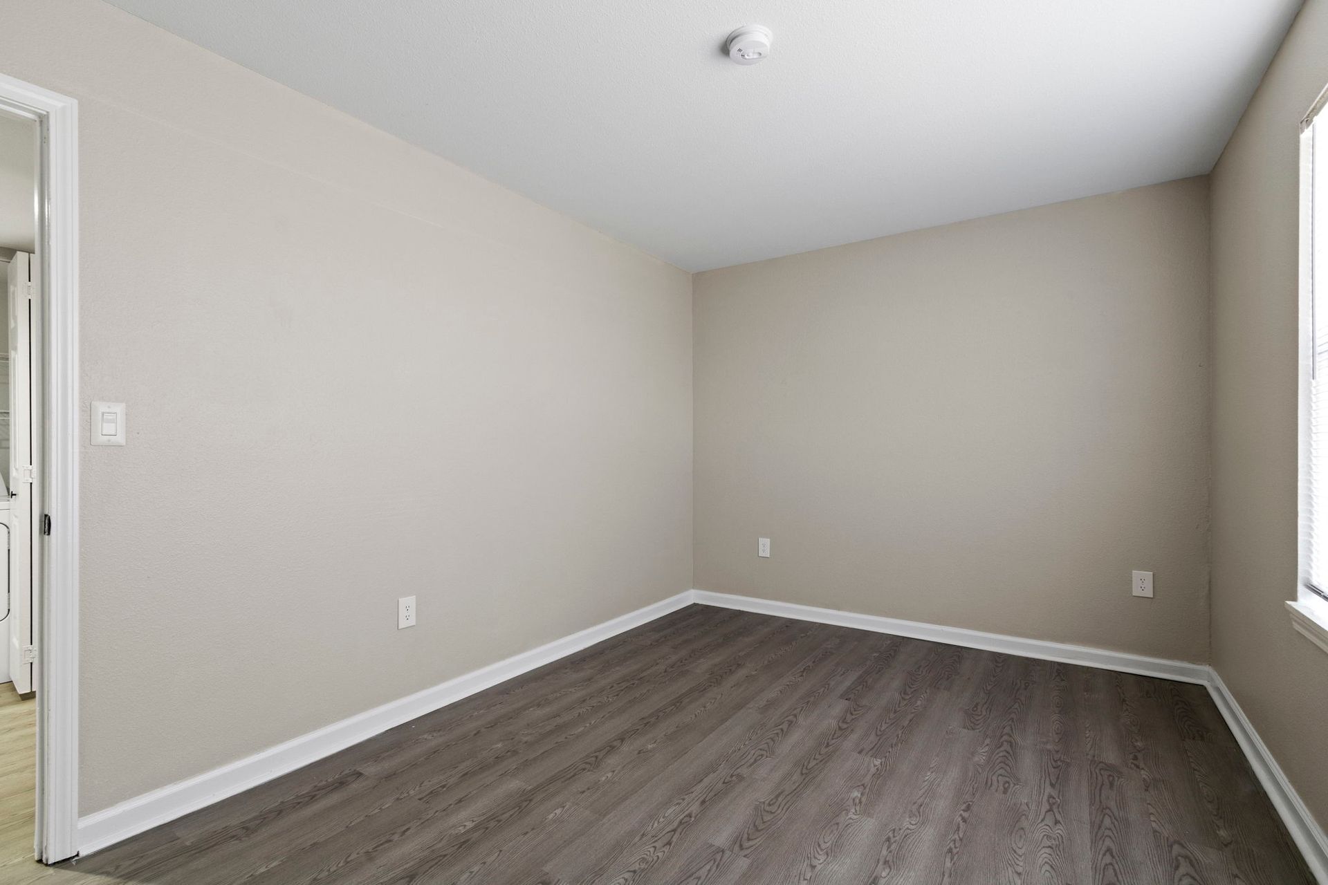 Empty room with light gray walls, dark wood-look floor, and a window on the right.