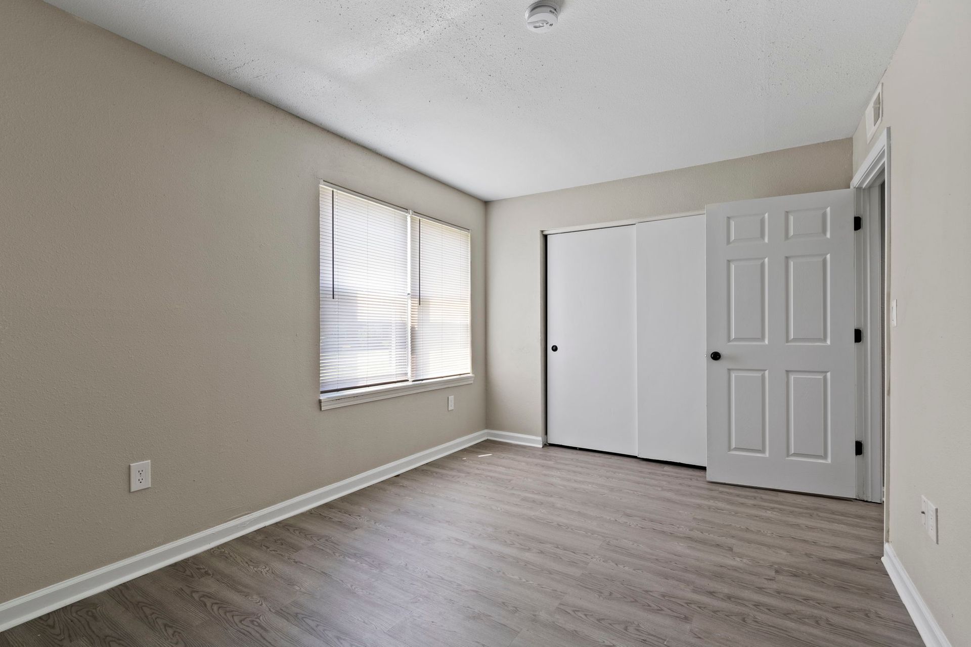 Empty bedroom with grey walls, wood-look floor, window with blinds, closet, and white door.