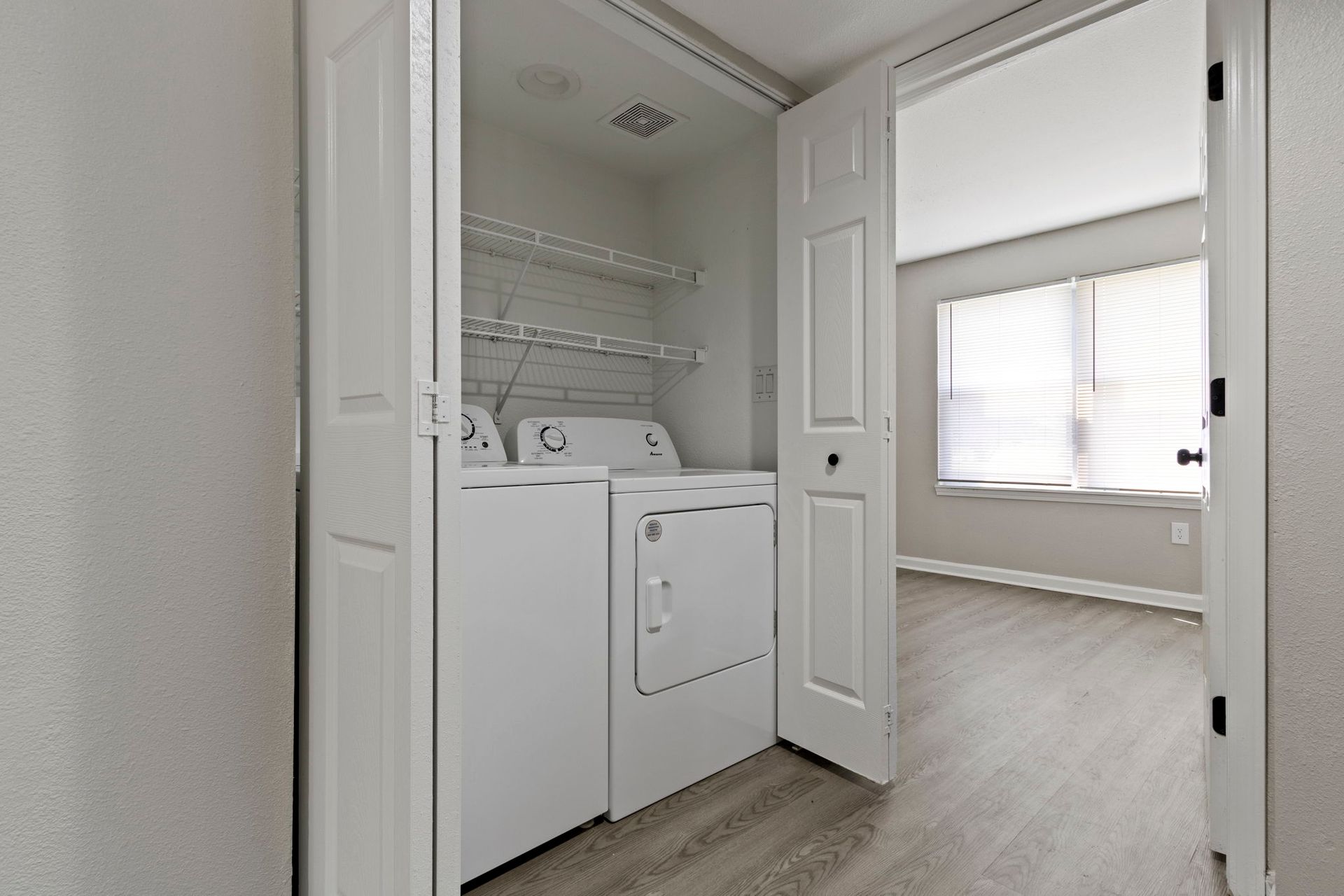 White washer and dryer in a closet with folding doors, next to a room with a window.