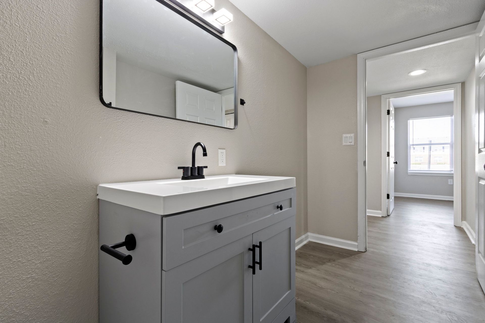 Gray vanity in a bathroom with a black faucet and mirror; doorway to a hallway with a window.