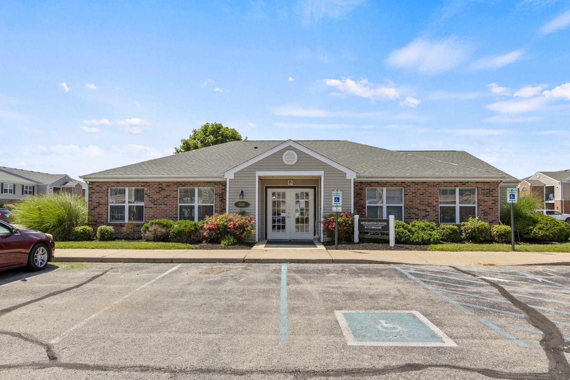 Apartment clubhouse exterior with parking. Brick facade, double doors, blue sky, accessible parking space.