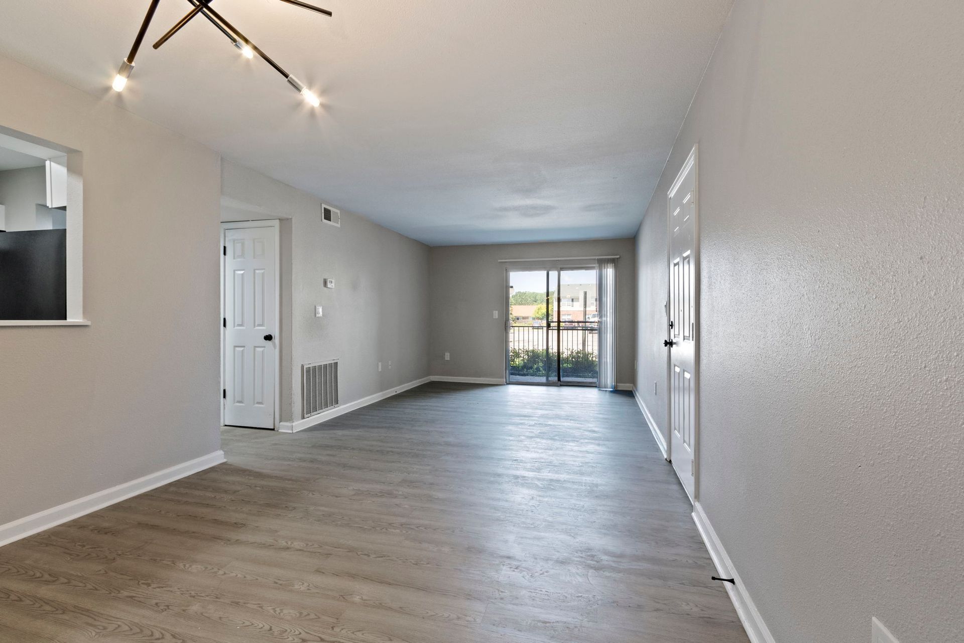 Empty apartment with light gray walls, wood-look flooring, and sliding glass door to balcony.