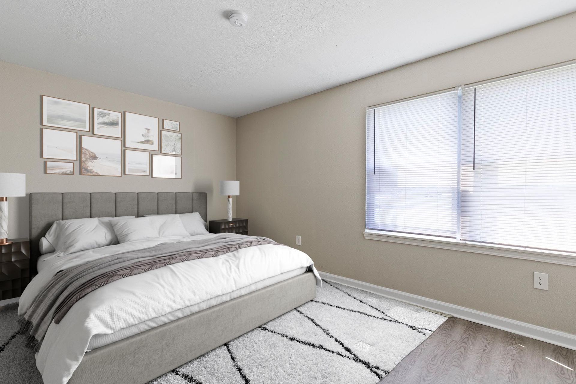 Bedroom with bed, art, rug, and window with blinds. Beige walls, white bedding, and natural light.